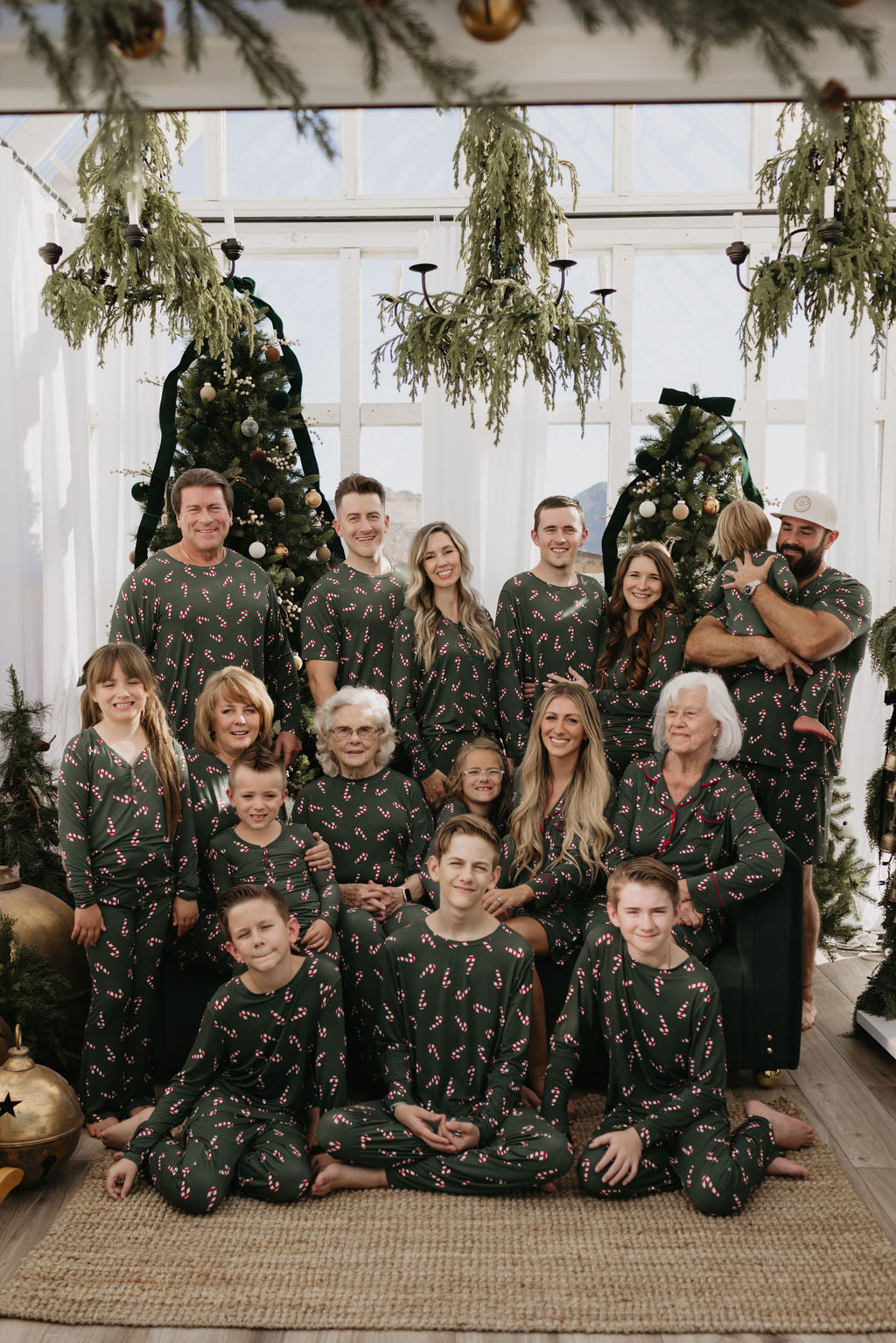 A large family in matching pajamas with candy cane patterns poses in front of Christmas trees inside a festive, plant-decorated room. Some stand, others sit, including children and elderly members, all smiling warmly.