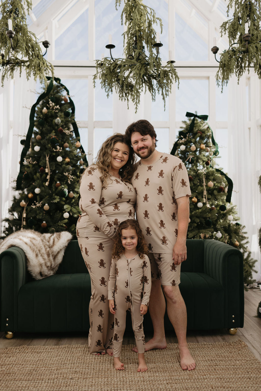 A smiling family wearing matching gingerbread-themed pajamas poses in front of a festive scene with two decorated Christmas trees and a green sofa.