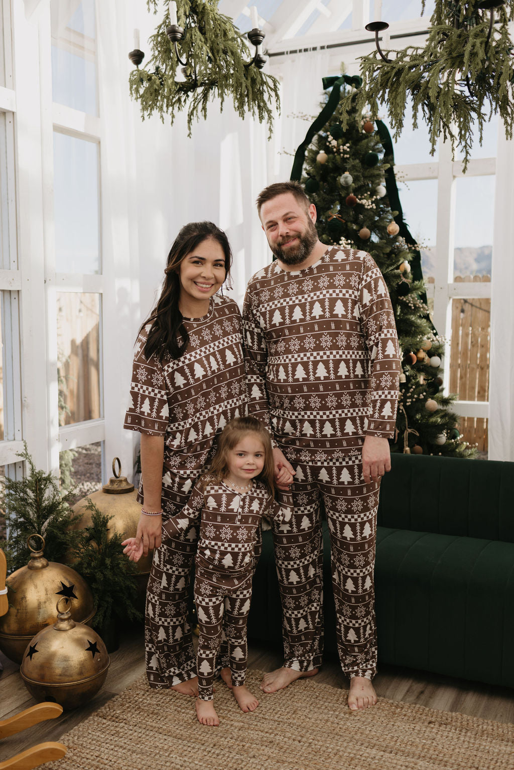 A family of three in matching Christmas pajamas smiles in a festive living room with a decorated tree. Greenery and large ornaments enhance the cozy holiday atmosphere.