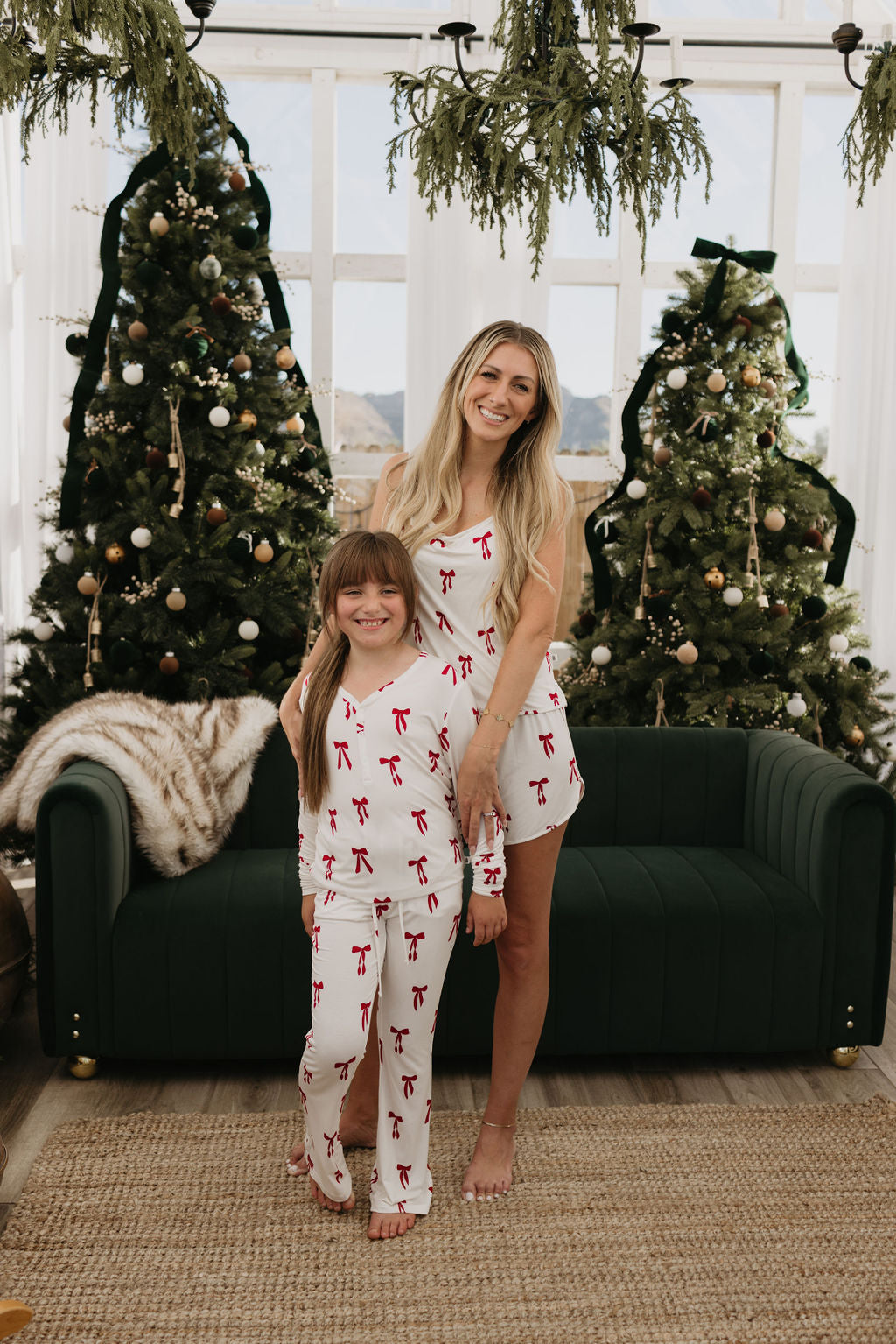 A woman and child wearing matching candy cane pajamas smile in front of a green couch. Two decorated Christmas trees stand in the background, and greenery hangs from the ceiling.