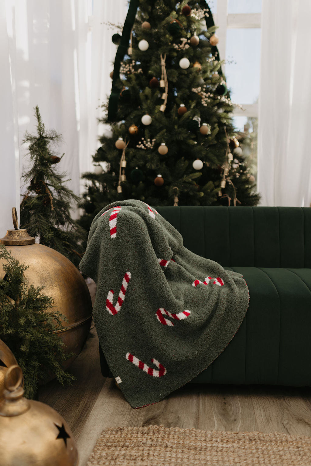 A green couch draped with a blanket featuring candy cane patterns is set in front of a decorated Christmas tree. Two large gold ornaments rest nearby, with greenery enhancing the festive atmosphere.