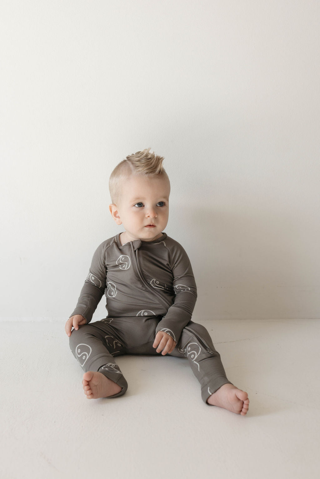 A baby with short blond hair sits barefoot against a white wall, wearing a long-sleeve, gray onesie with a white abstract pattern.