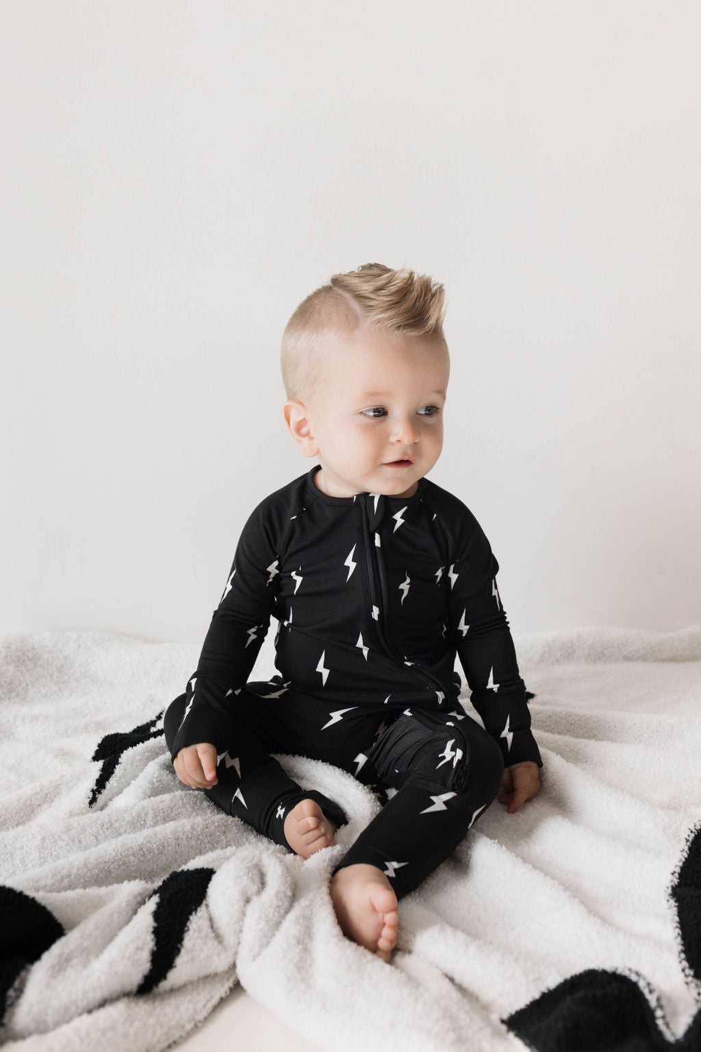 A baby with short blond hair sits on a white blanket, wearing a black outfit with white lightning bolt patterns, looking slightly to the side.