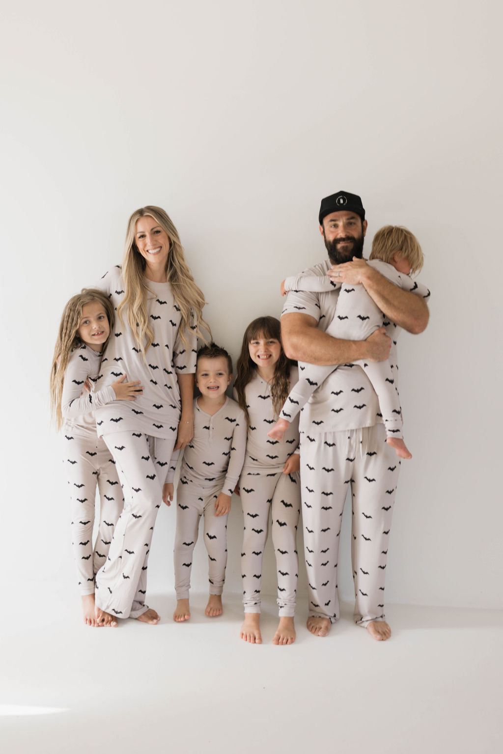 A family of six poses in matching white pajamas with black bat patterns. They stand barefoot against a plain background, smiling and embracing each other.