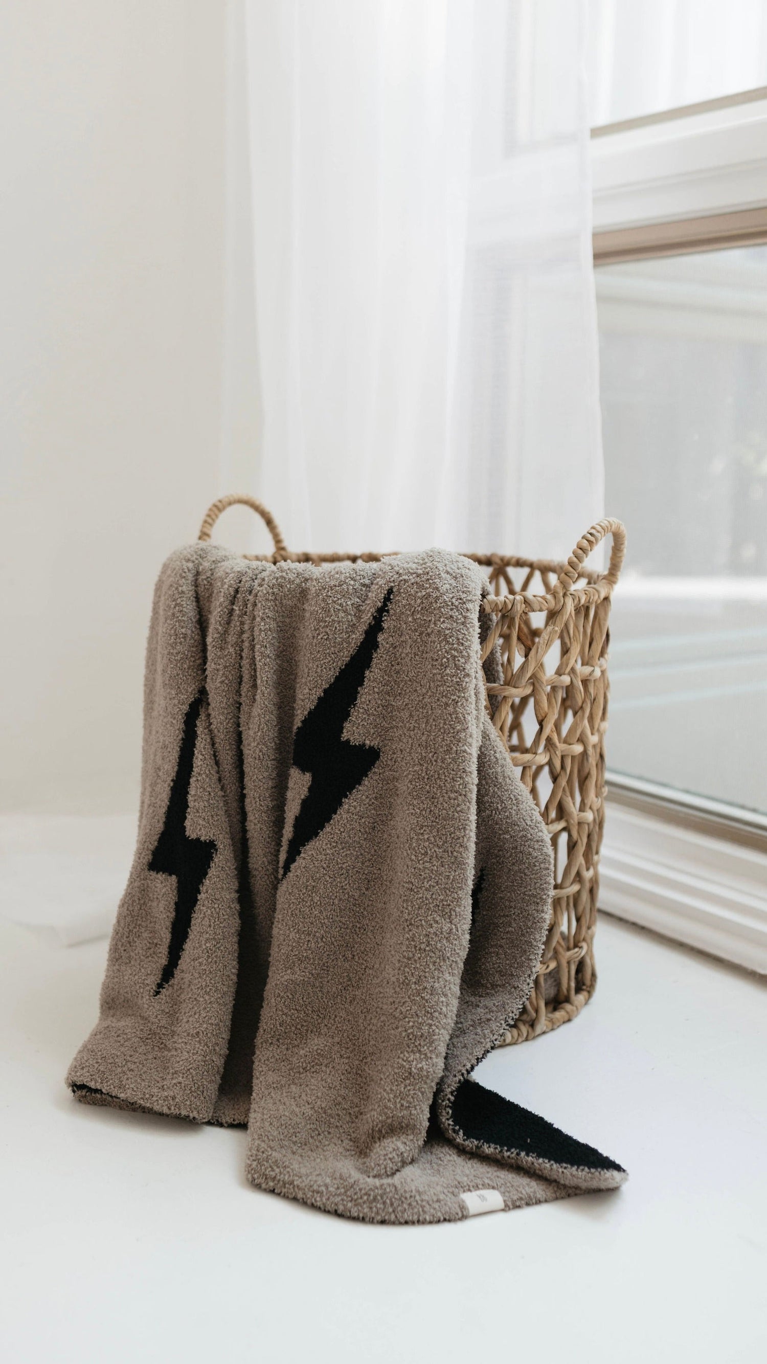 A soft, beige towel with black lightning bolt designs draped over a woven basket near a window with sheer white curtains.