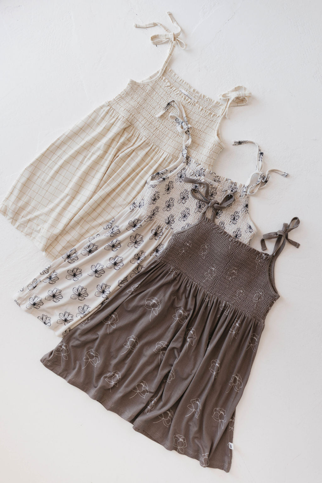 Three sleeveless sundresses with shoulder ties are laid out. The first is beige with checks, the second is white with floral patterns, and the third is dark brown with a floral print.