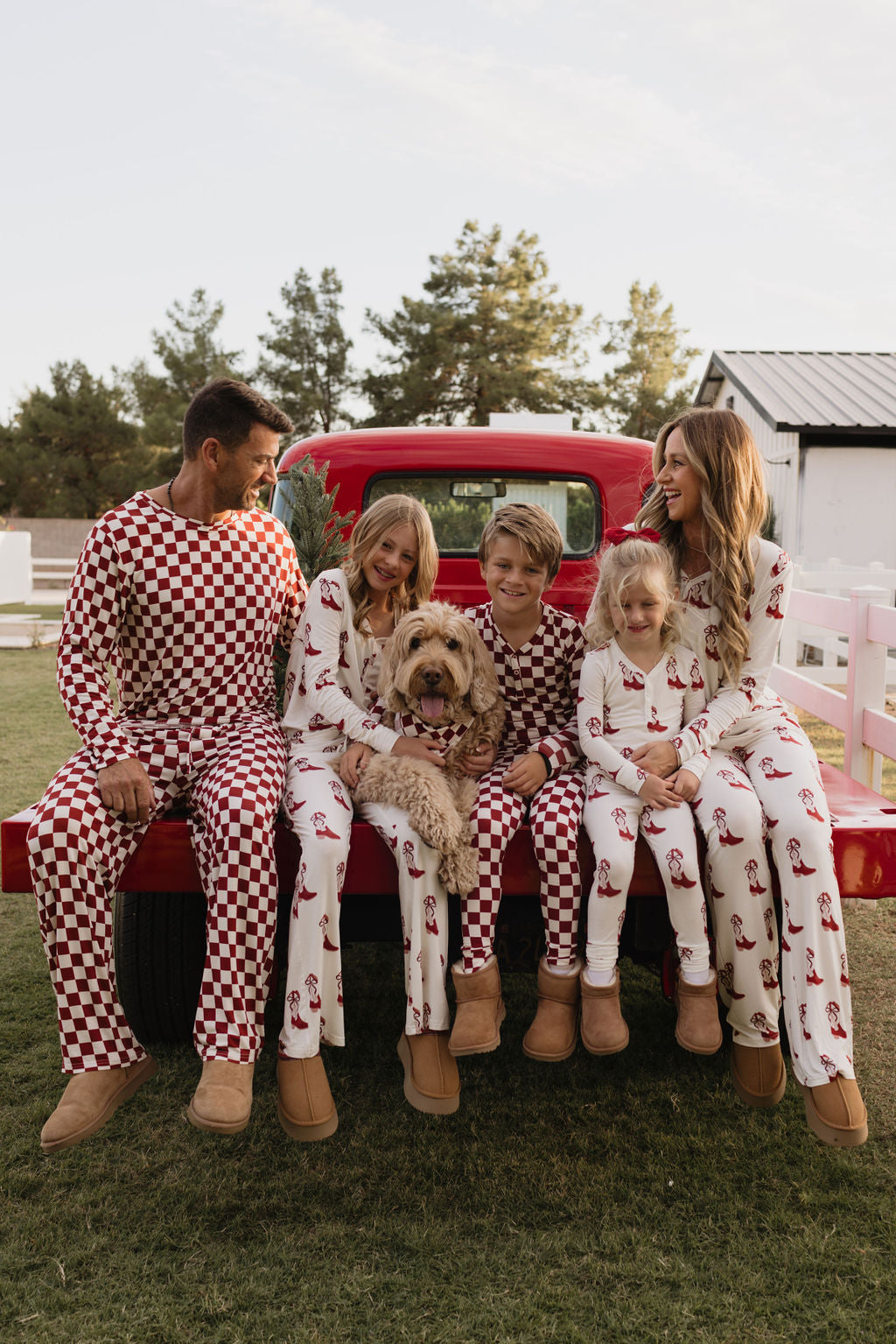 A family and a dog sit on the back of a red truck. They're wearing matching pajamas with red and white patterns. The background features a barn and trees under a clear sky.