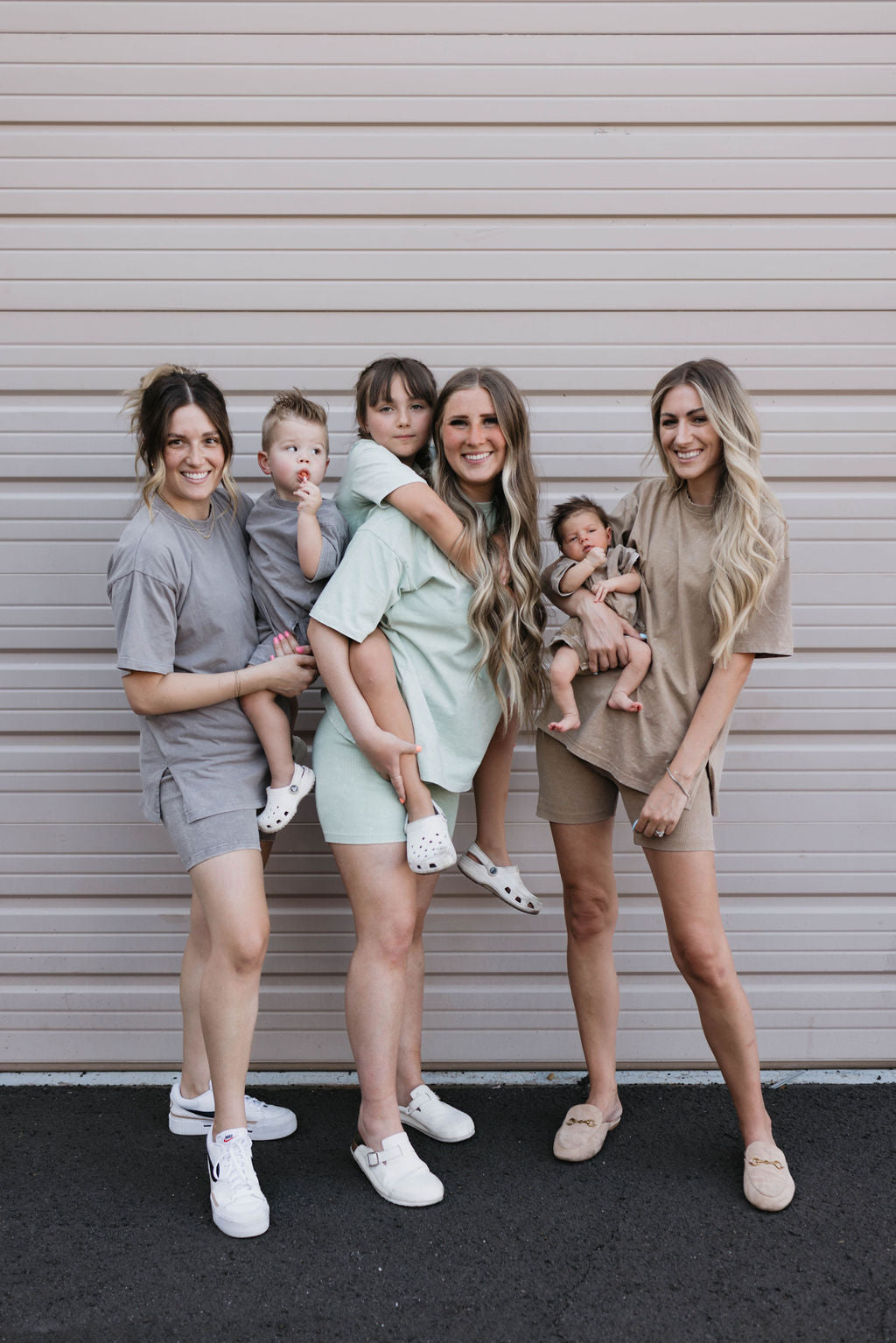 Three women and three children stand smiling in front of a garage door. Two women hold infants while one child is carried piggyback. All are dressed in casual, neutral-colored outfits.