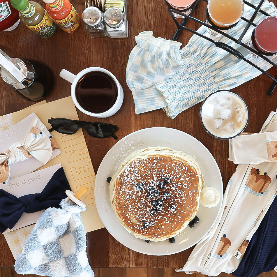 A plate of pancakes with blueberries and powdered sugar sits on a wooden table with coffee, juice, syrup, utensils, napkins, bow ties, sunglasses, and condiments.