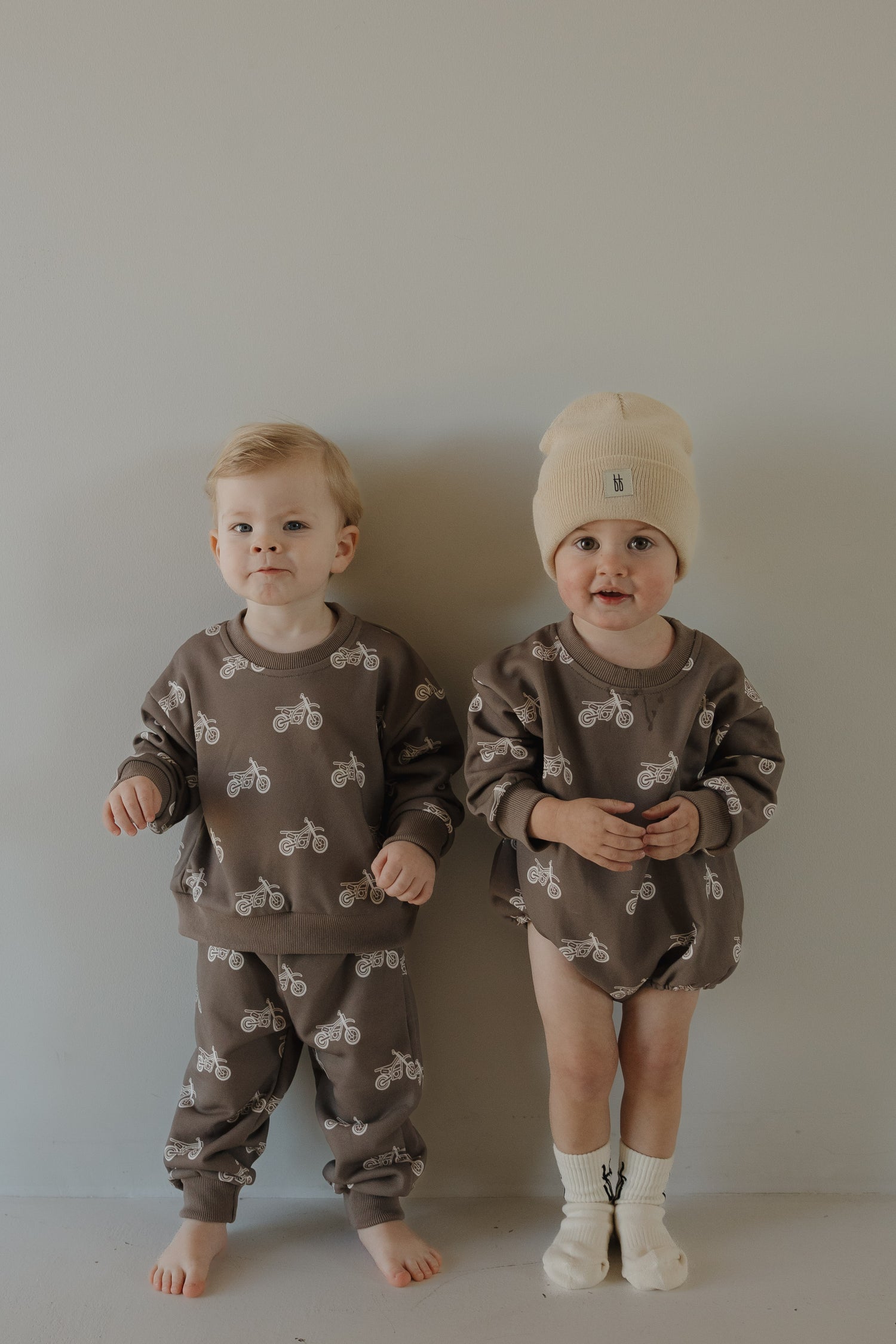 Two young children stand against a plain wall wearing matching brown outfits with bicycle prints. One child wears pants, the other a beanie and shorts. Both are barefoot, and one wears socks.