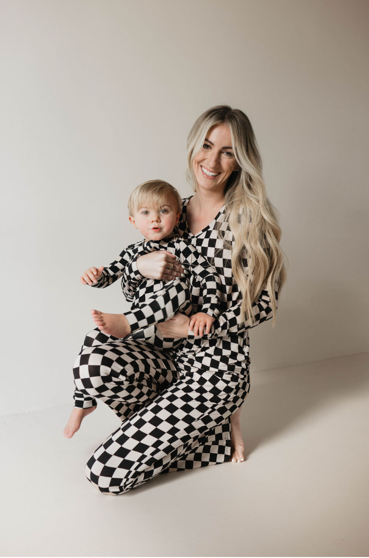 A woman with long blonde hair is kneeling, holding a young child. Both are wearing matching black and white checkered outfits. The background is plain and light-colored, creating a calm setting.