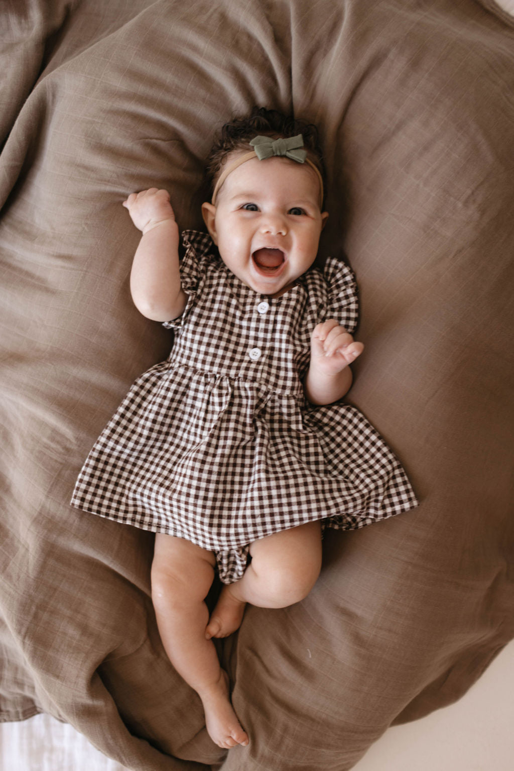 A smiling baby is lying on a brown blanket, wearing a checkered dress and a bow headband. The baby has one hand raised and looks joyful.