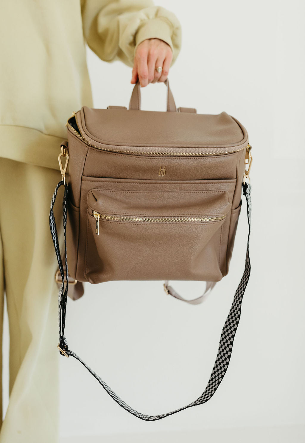 Person holding a taupe-colored bag with a top handle and detachable strap. The bag features multiple zippers and a textured strap detail. The background is plain and the person is wearing light-colored clothing.