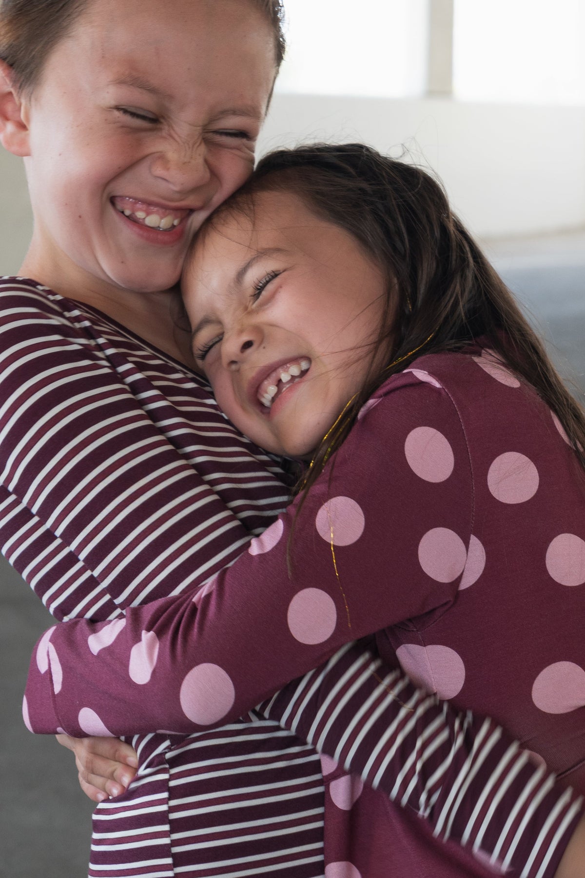 Two young girls hug tightly, smiling with eyes closed. One wears a maroon and white striped dress, the other wears a maroon dress with large pink polka dots. They appear joyful and happy.