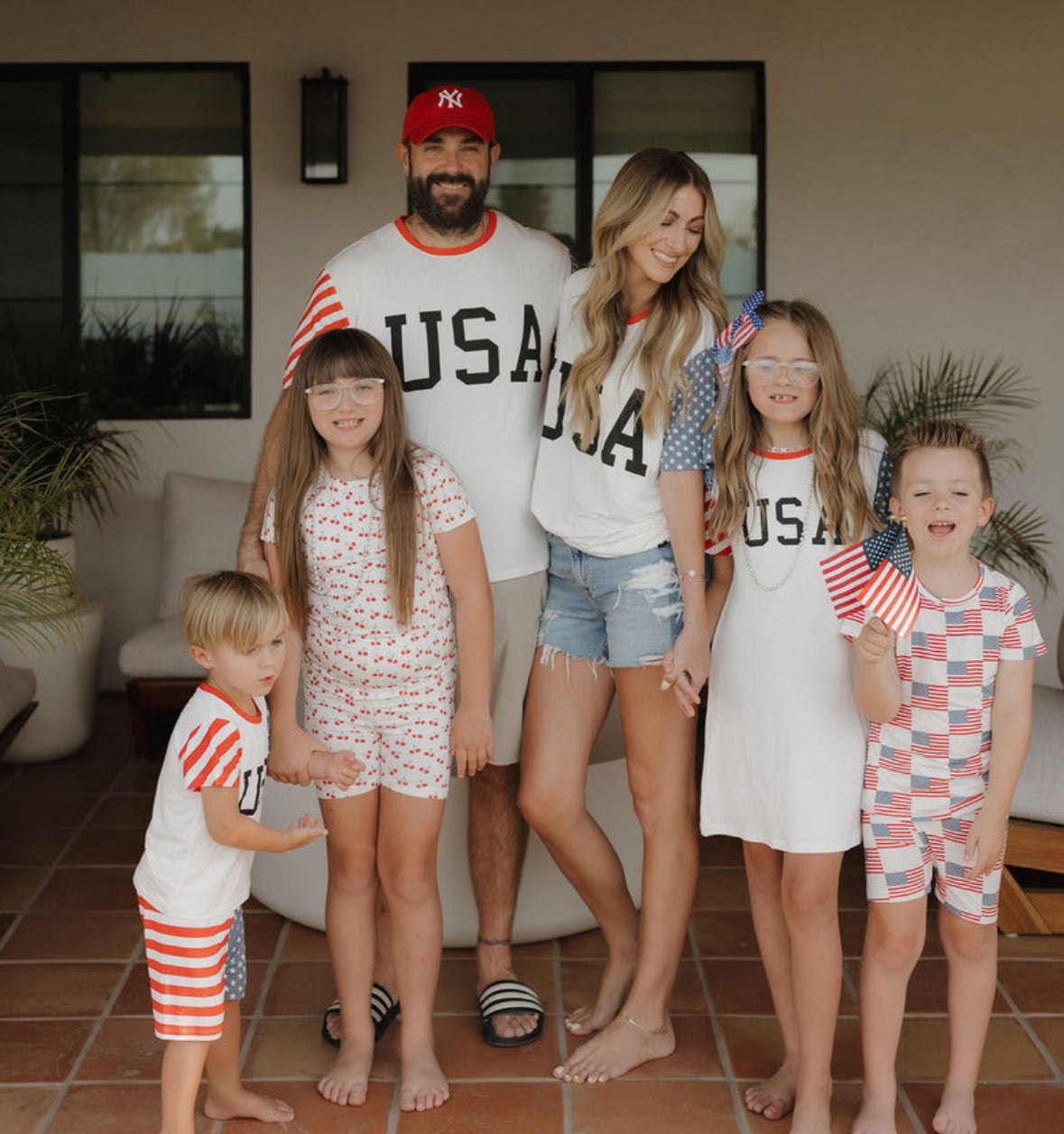 A family of five stands outside, smiling and holding small American flags. The parents and children wear red, white, and blue outfits with "USA" and flag patterns, celebrating a patriotic occasion.