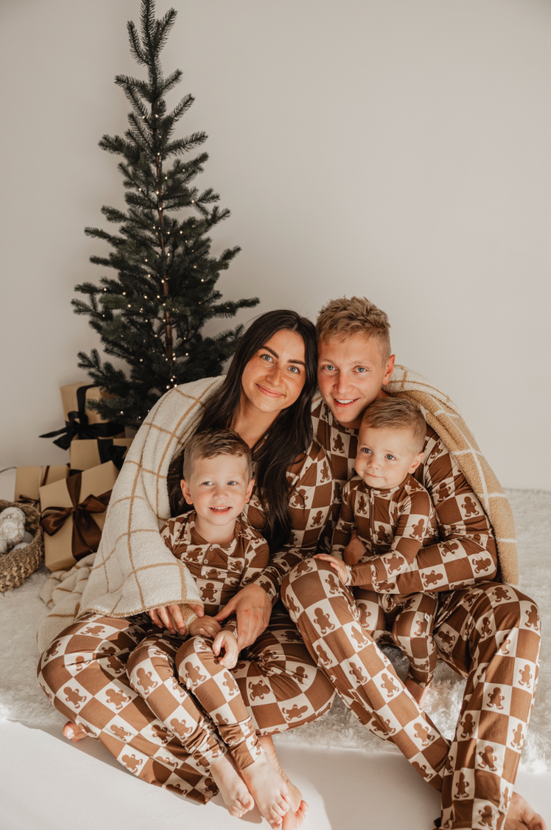 A family of four sits together in matching brown pajamas with a festive pattern, smiling and wrapped in a blanket. A decorated Christmas tree and gifts are in the background.