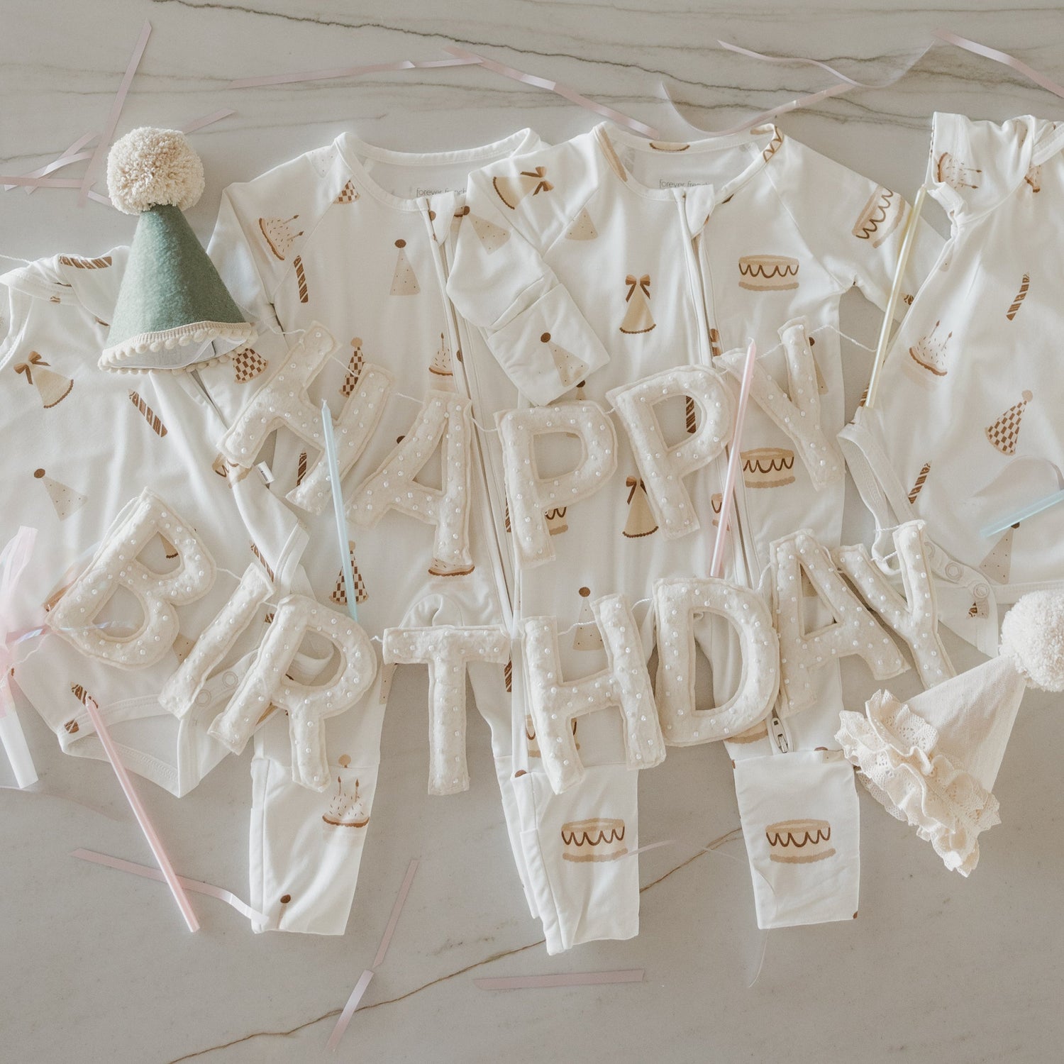 Five white baby onesies with cake prints are arranged on a table with pastel ribbons, a felt party hat, a lace hat, and "HAPPY BIRTHDAY" spelled out in plush letters on top.