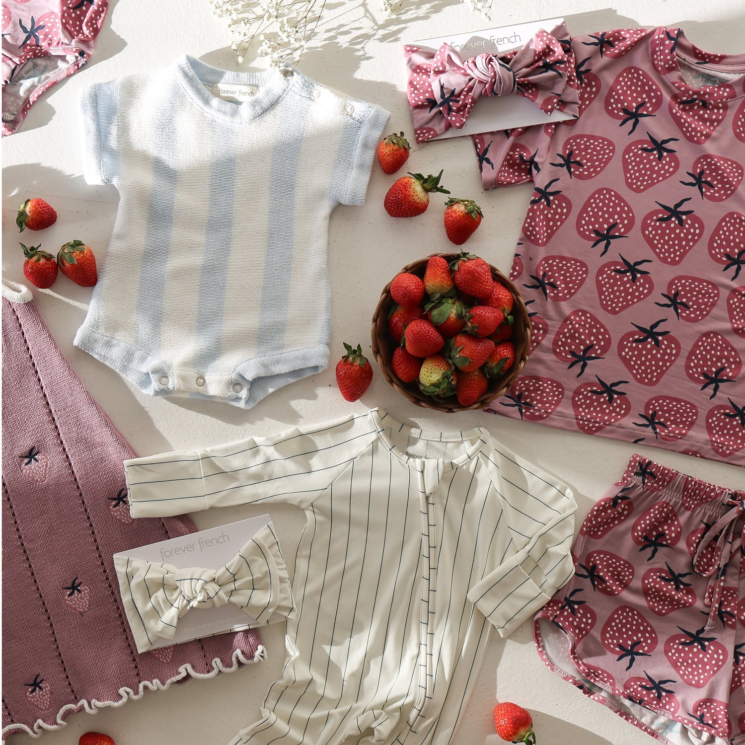 Flat lay of baby clothes, including a striped onesie, blue and white bodysuit, bows, a pink blanket with a scalloped edge, strawberry-patterned fabric, and a bowl of fresh strawberries.