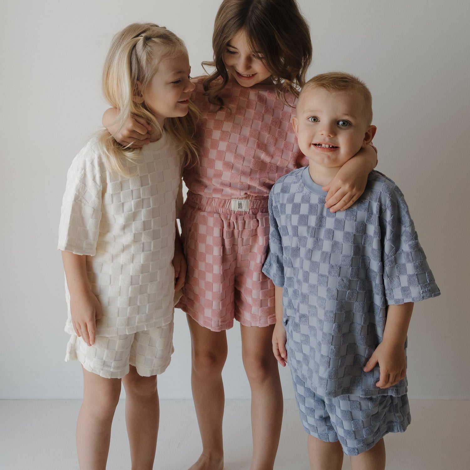 Three young children stand together smiling, wearing matching textured outfits in cream, pink, and blue, against a plain white background.