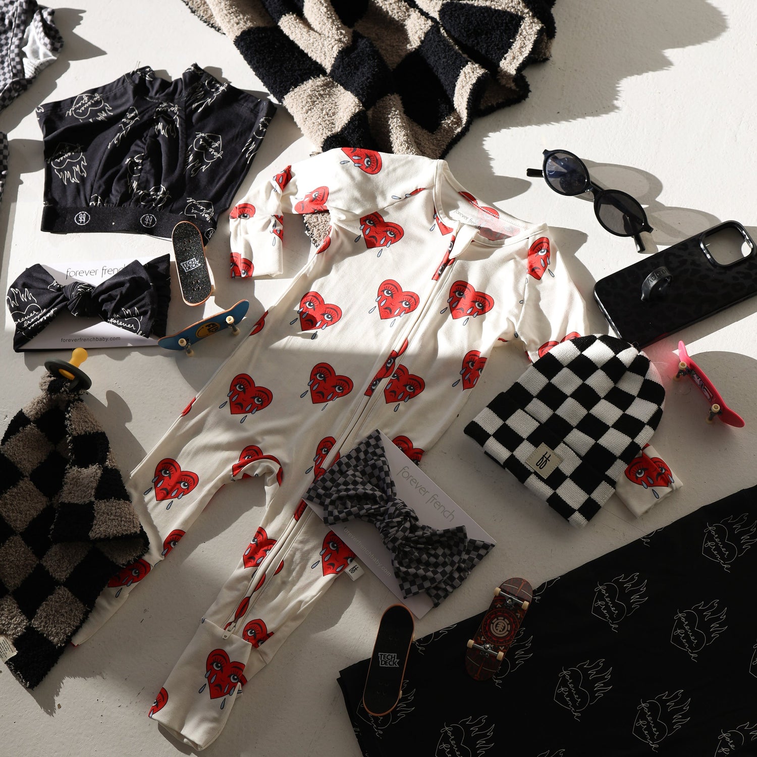 Flat lay of baby clothes, including a white onesie with red heart faces, a black printed shirt, headbands, a knit hat, sunglasses, hot water bottles, books, and accessories on a white surface.