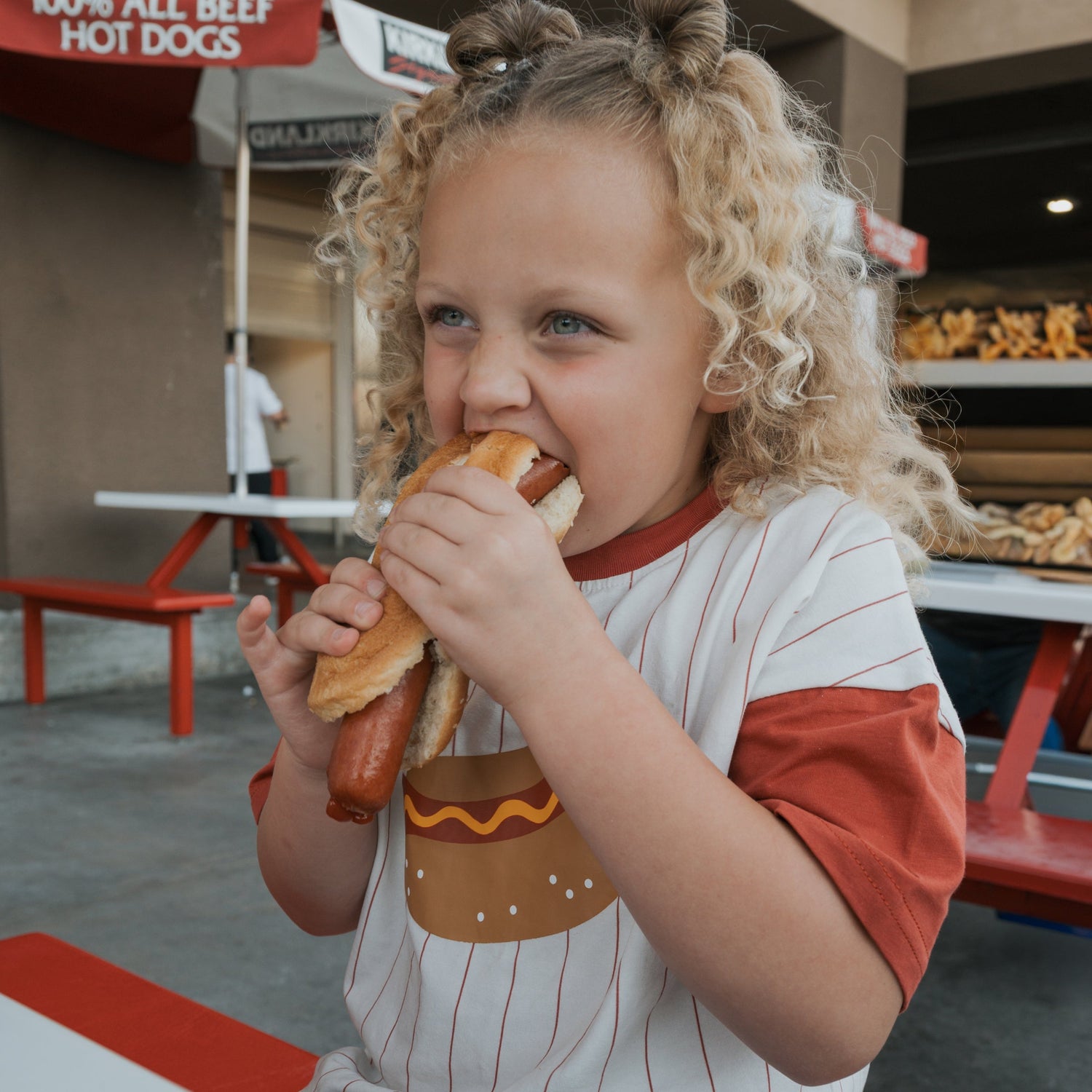 A young child with curly blonde hair is sitting at a red and white table, smiling and taking a big bite of a hot dog. There are hot dog signs and food stands in the background.