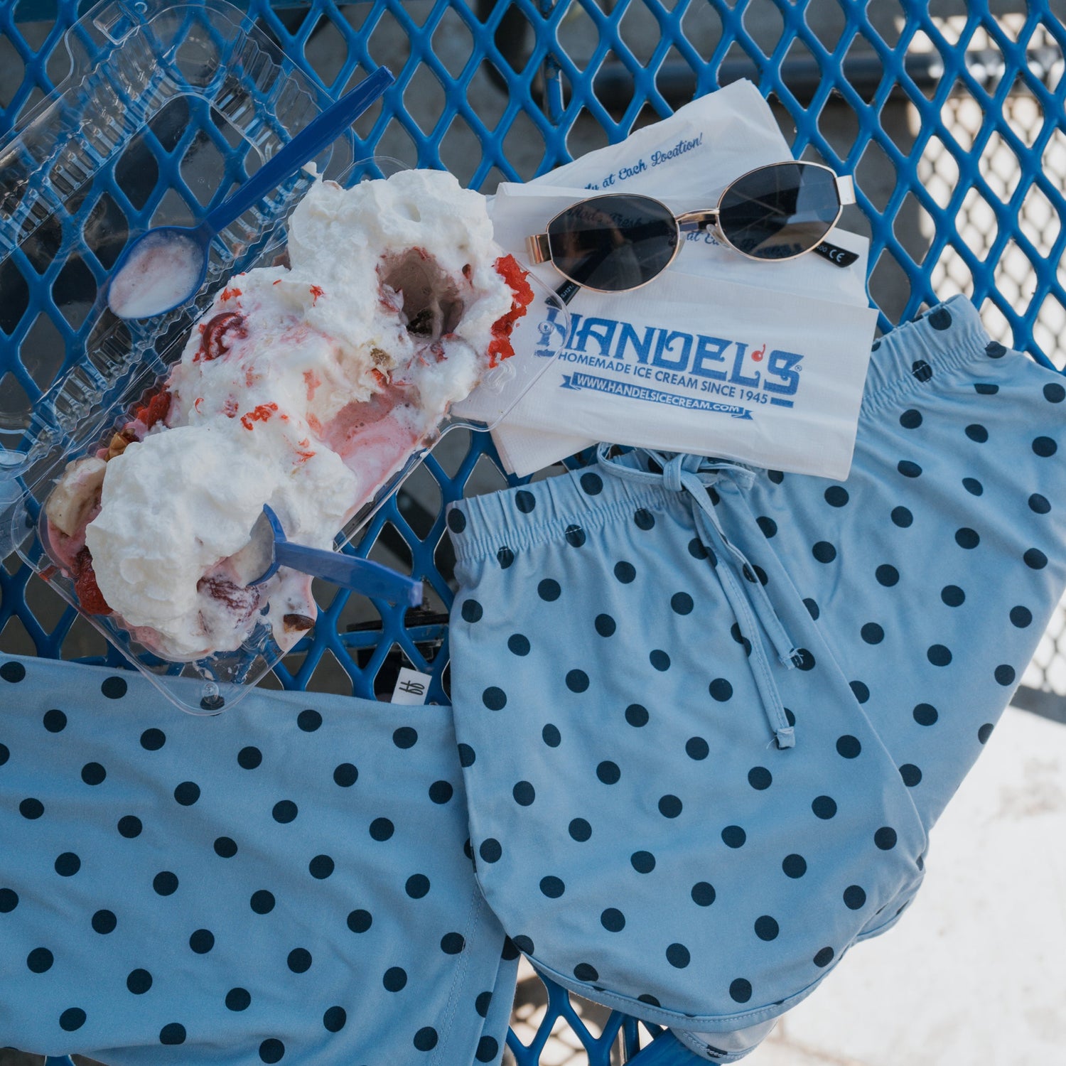 A banana split with whipped cream in a clear container, sunglasses, napkins, and blue polka dot shorts on a blue metal table in sunlight.