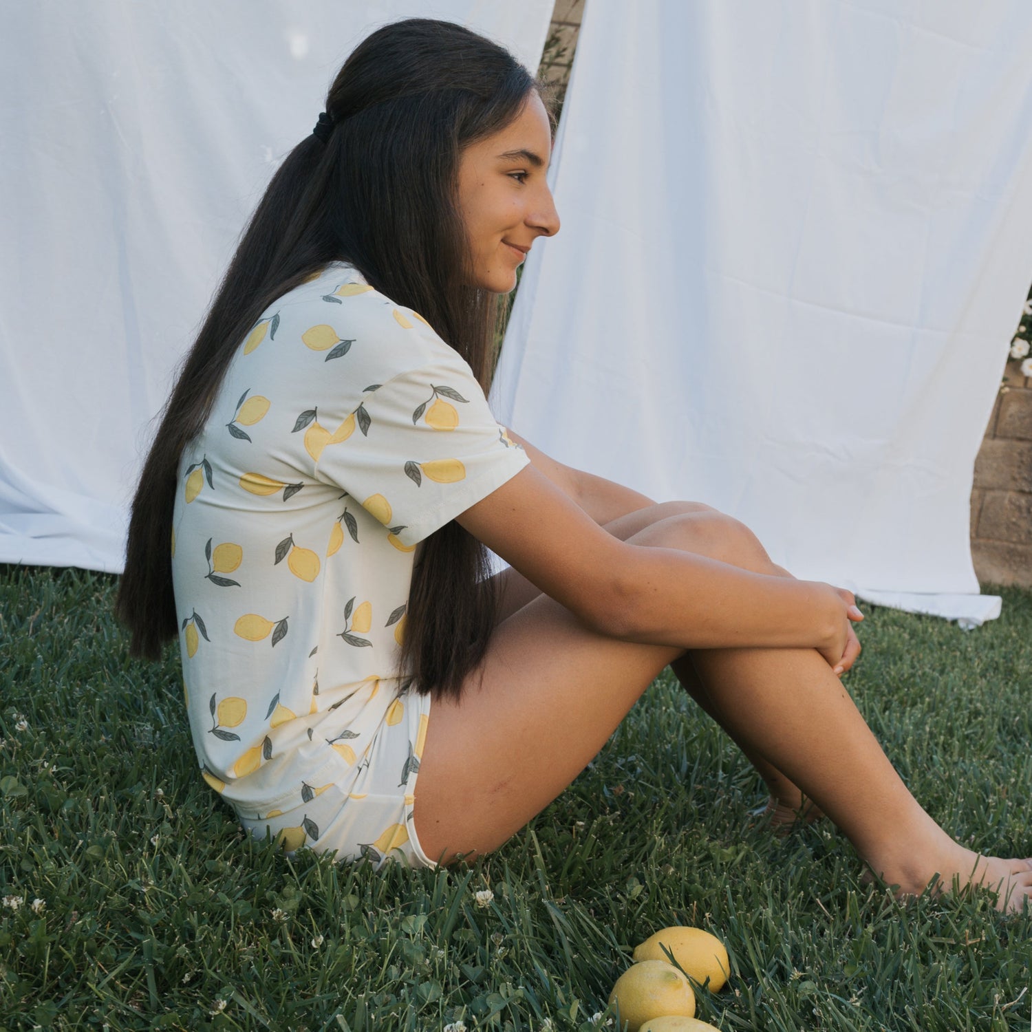 A person with long dark hair sits on green grass, wearing lemon-patterned pajamas. Three lemons are on the grass nearby, and white sheets hang in the background.