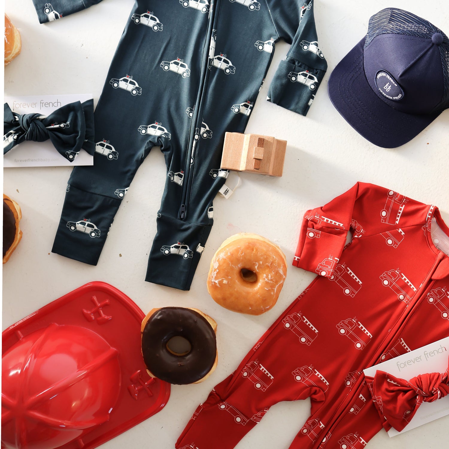 Flat lay of two baby onesies, one navy with white car prints and one red with white fire truck prints. Surrounding them are donuts, a bow tie, a toy fire hat, a toy block, and a blue cap.
