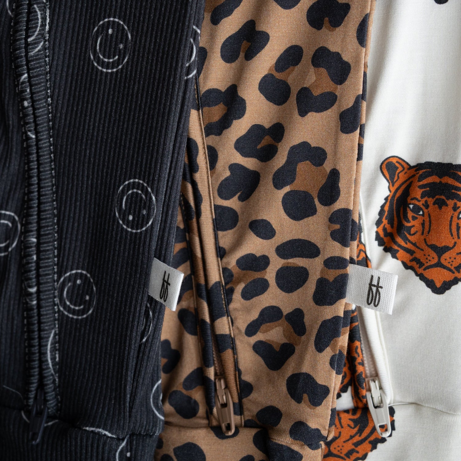 Three zippered garments are shown: one black with white smiley faces, one tan with black leopard spots, and one white with a tiger face. All have small white tags with “bb” on them.