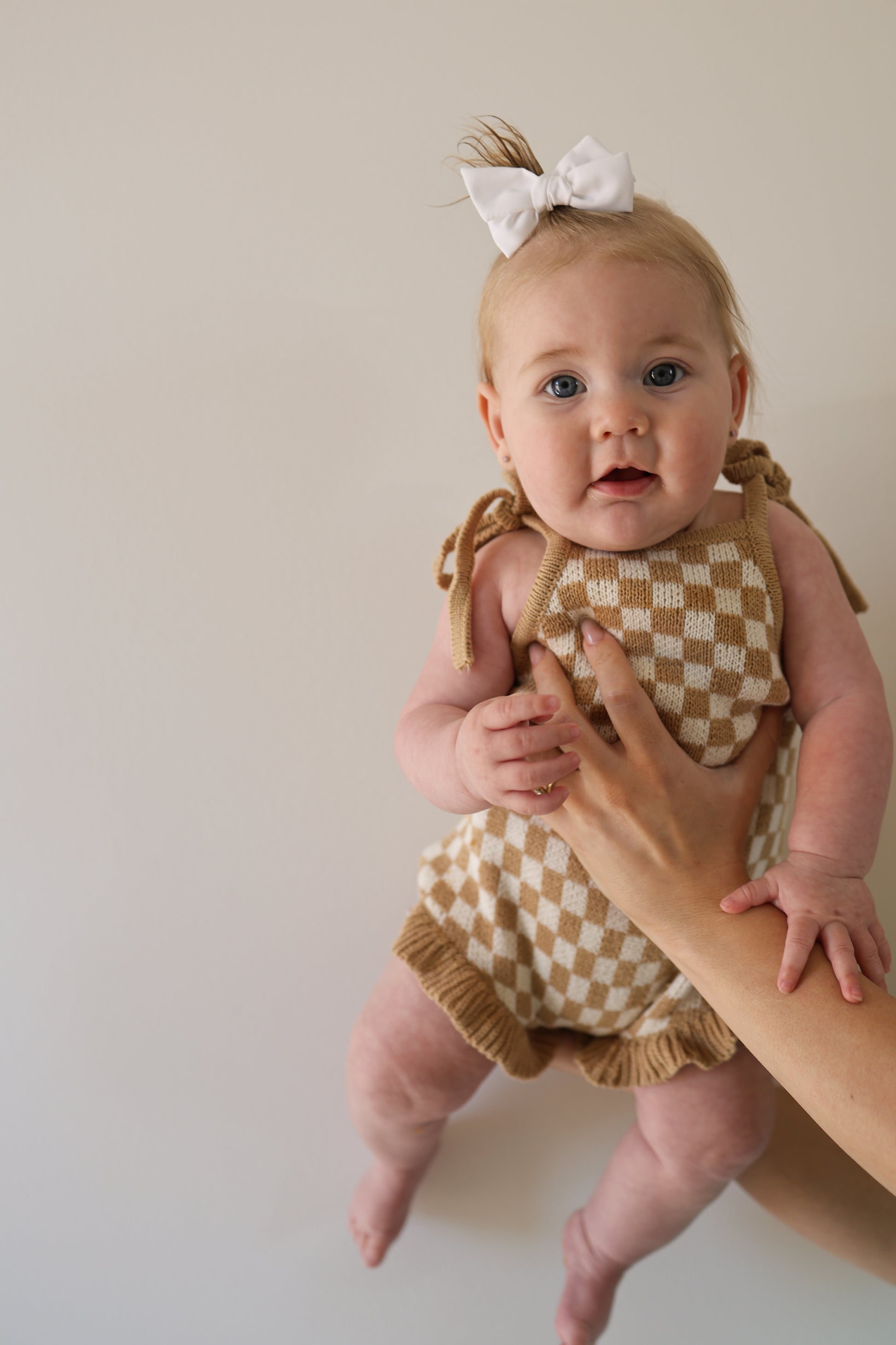 A baby wearing the forever french baby Knit Ruffle Romper in Latte Check with a white bow is held up by an adult against a plain light background.