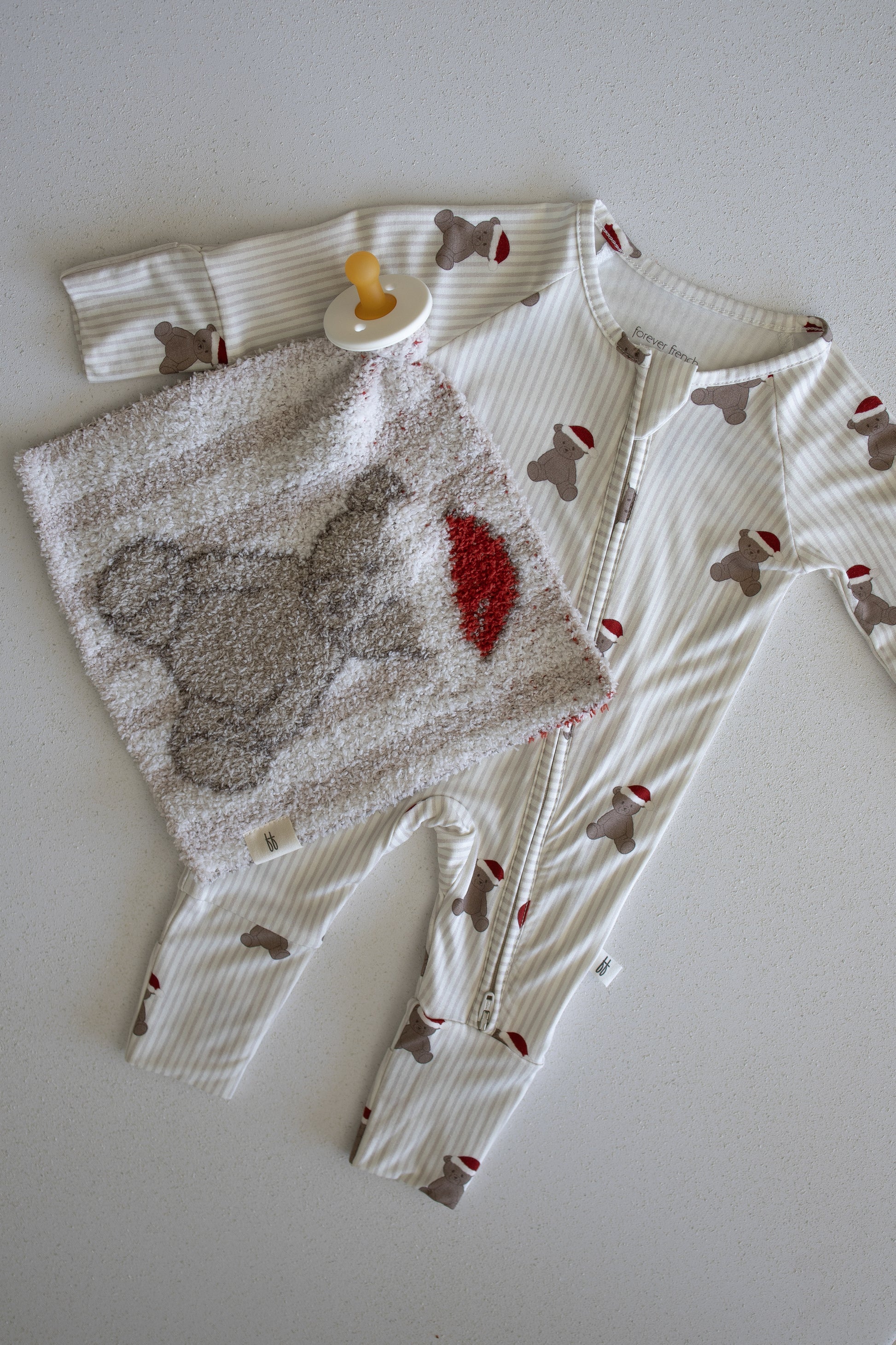 A Lovey | Santa Teddy from forever french baby—a soft, textured blanket featuring an adorable bear and red heart—is arranged alongside a cozy yellow pacifier and bear-print onesie on a light surface, making it perfect for your little one's cuddly moments with their favorite lovey.
