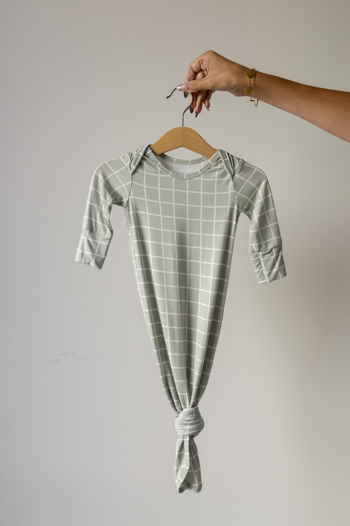 A hand holds the forever french baby Bamboo Knotted Gown | Foggy Grid—a long-sleeve, light gray bamboo baby gown with a white grid pattern and bottom knot—against a plain light background, making it an ideal newborn essential for those seeking soft, hypoallergenic baby clothing.