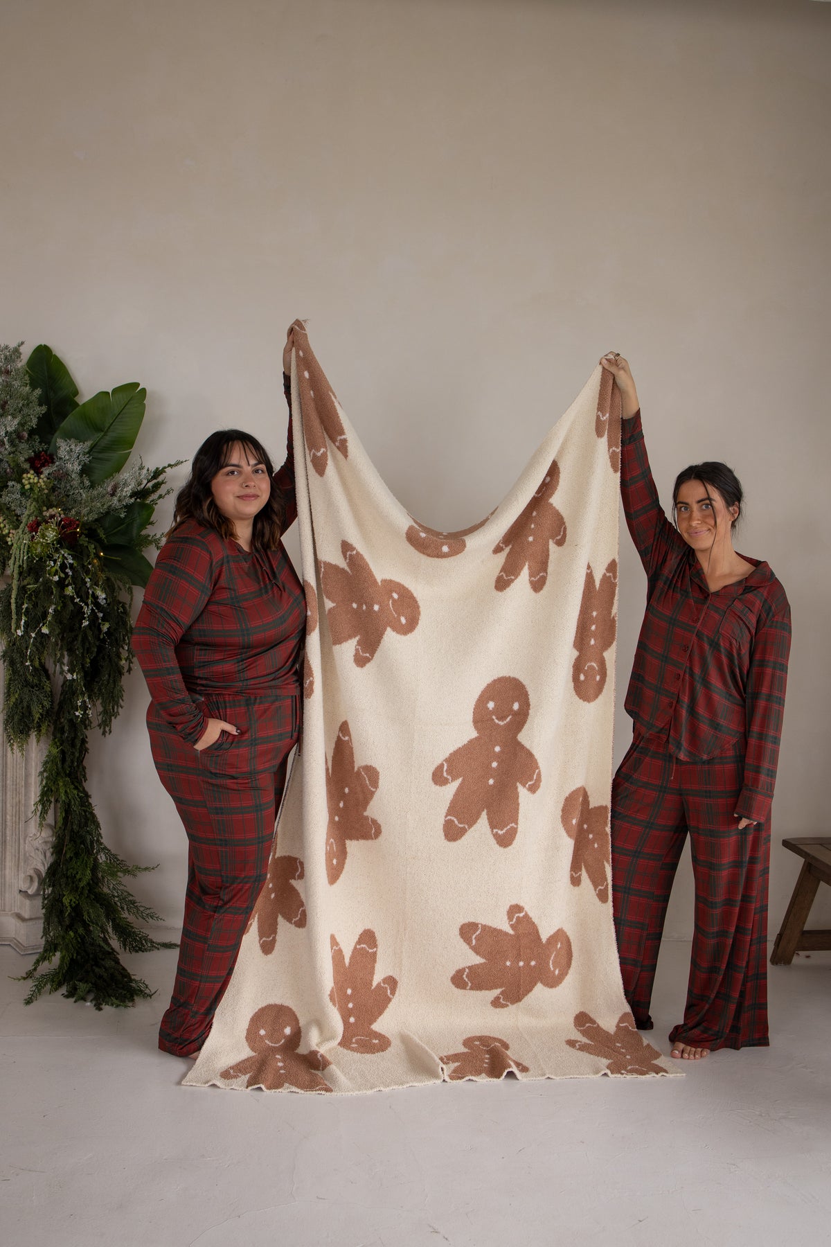 Two women in red plaid pajamas hold up a soft, brown gingerbread-patterned *PRE-ORDER* Plush Blanket | Gingersnap.
