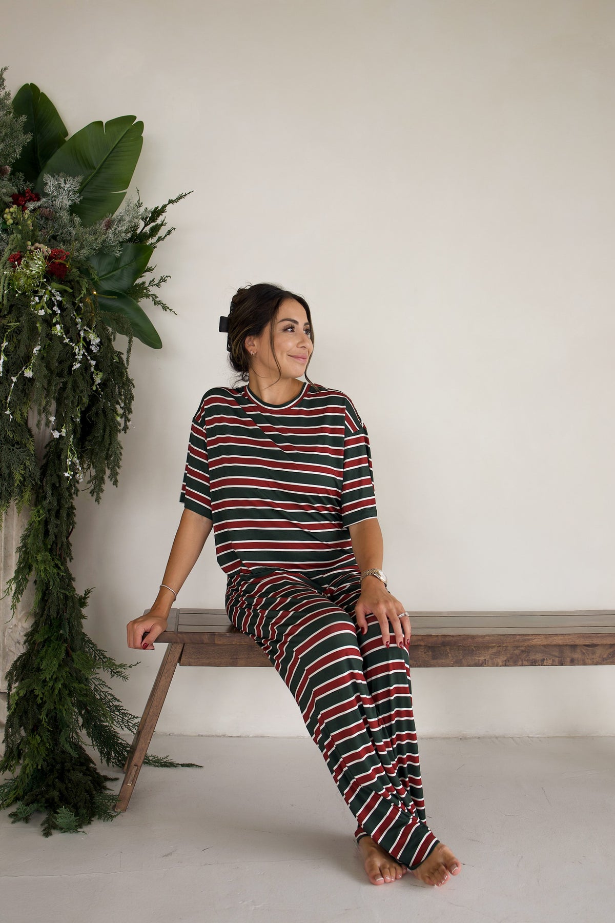 A woman wears the forever french baby Women’s Boxy Tee Lounge Set | Holiday Stripe, sitting on a wooden bench in a matching short-sleeved top and wide-leg lounge pants with horizontal red, green, and white stripes.