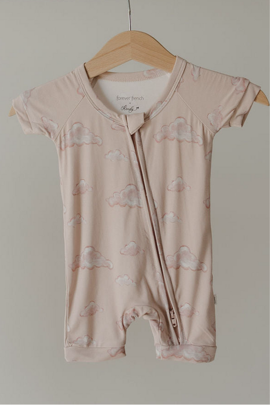The Kendy 3 Bamboo Zip Shortie | Ruby is a short-sleeved, light pink baby romper with a cloud pattern, made from premium bamboo pajamas material and featuring a diagonal zipper from collar to left leg. Shown on a wooden hanger.