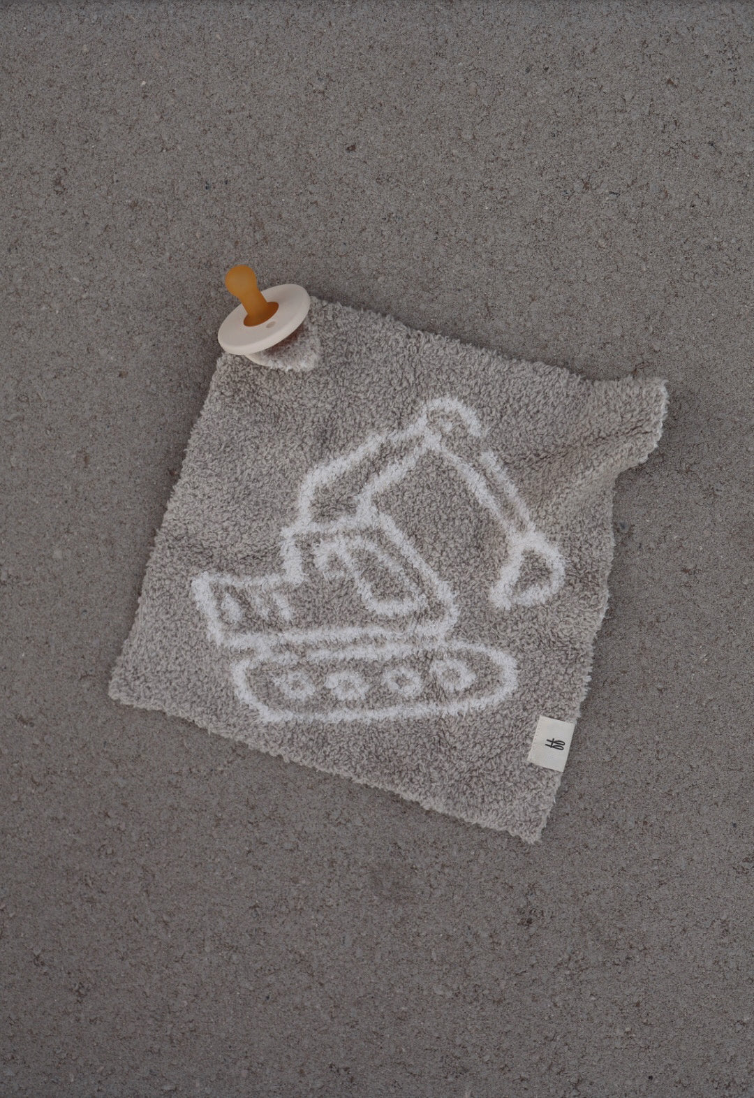 The Lovey | Excavator from forever french baby is a plush gray microfiber lovey with a white excavator outline and an attached brown pacifier, shown on a textured gray surface.