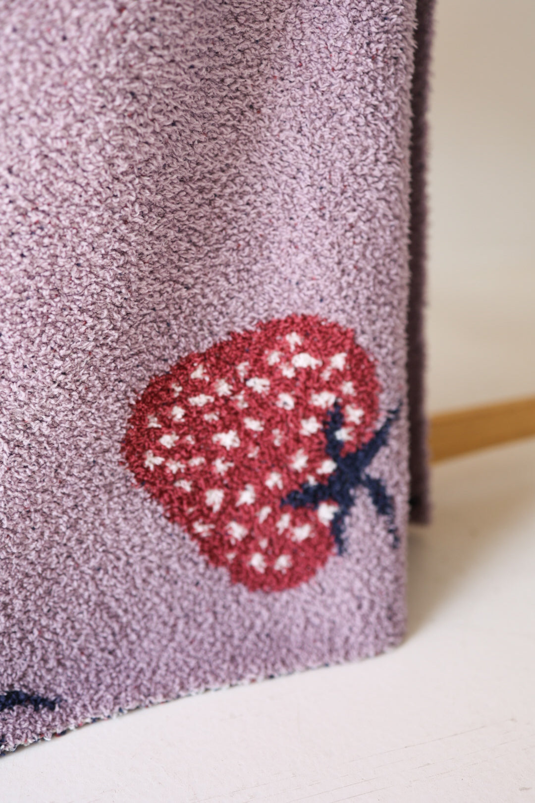 A close-up of the forever french baby Plush Blanket | Matilda in light purple velvet, featuring red strawberries with white seeds and dark stems. The fabric is draped, with a wooden rod partly visible in the background.