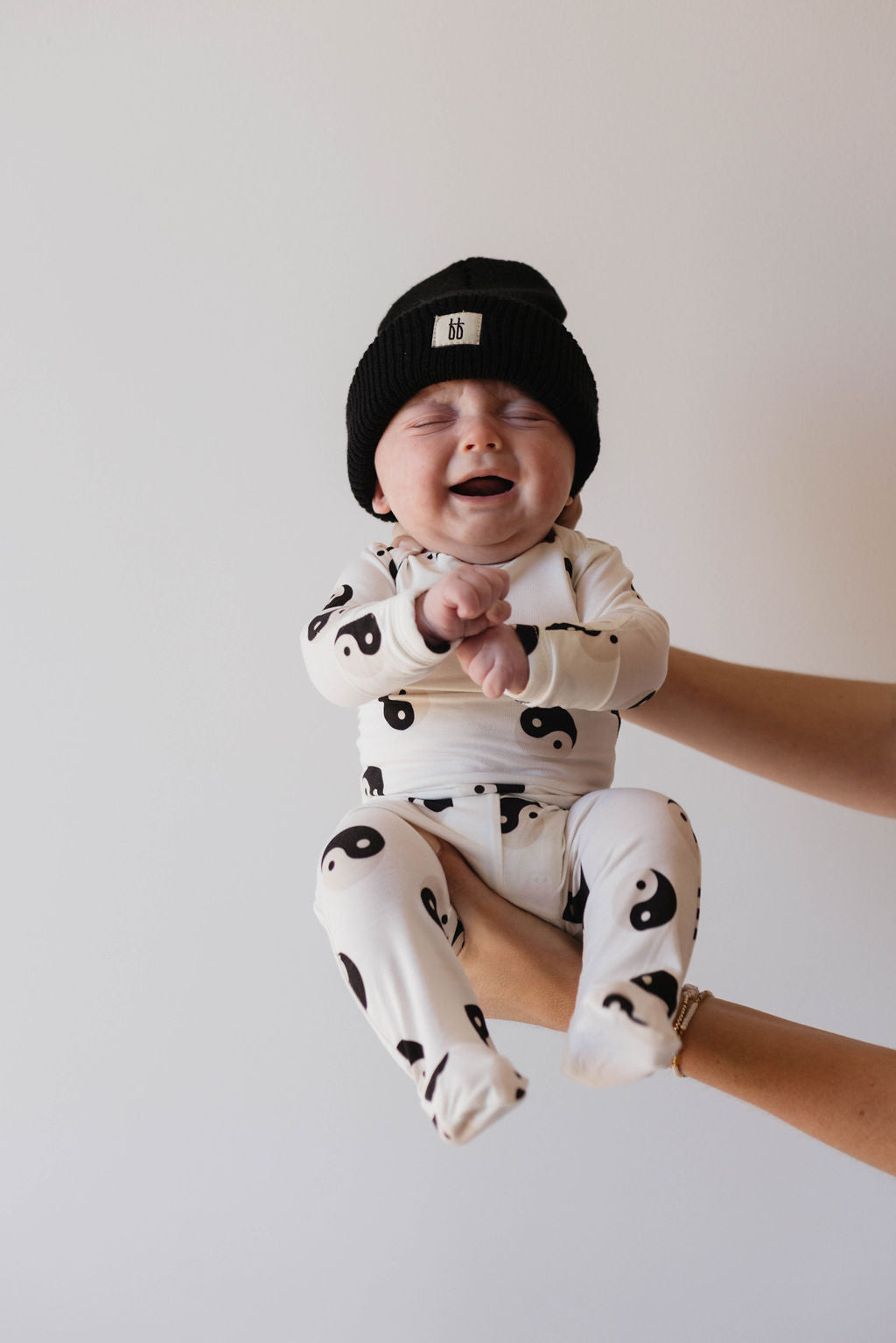 A baby sporting a beanie and outfit from the forever french baby Sleepy Time Set | Black & White Yin Yang, adorned with a yin-yang symbol pattern, is supported by adult hands against a plain background. The baby's eyes are closed and mouth open, giving the appearance of laughing or crying.