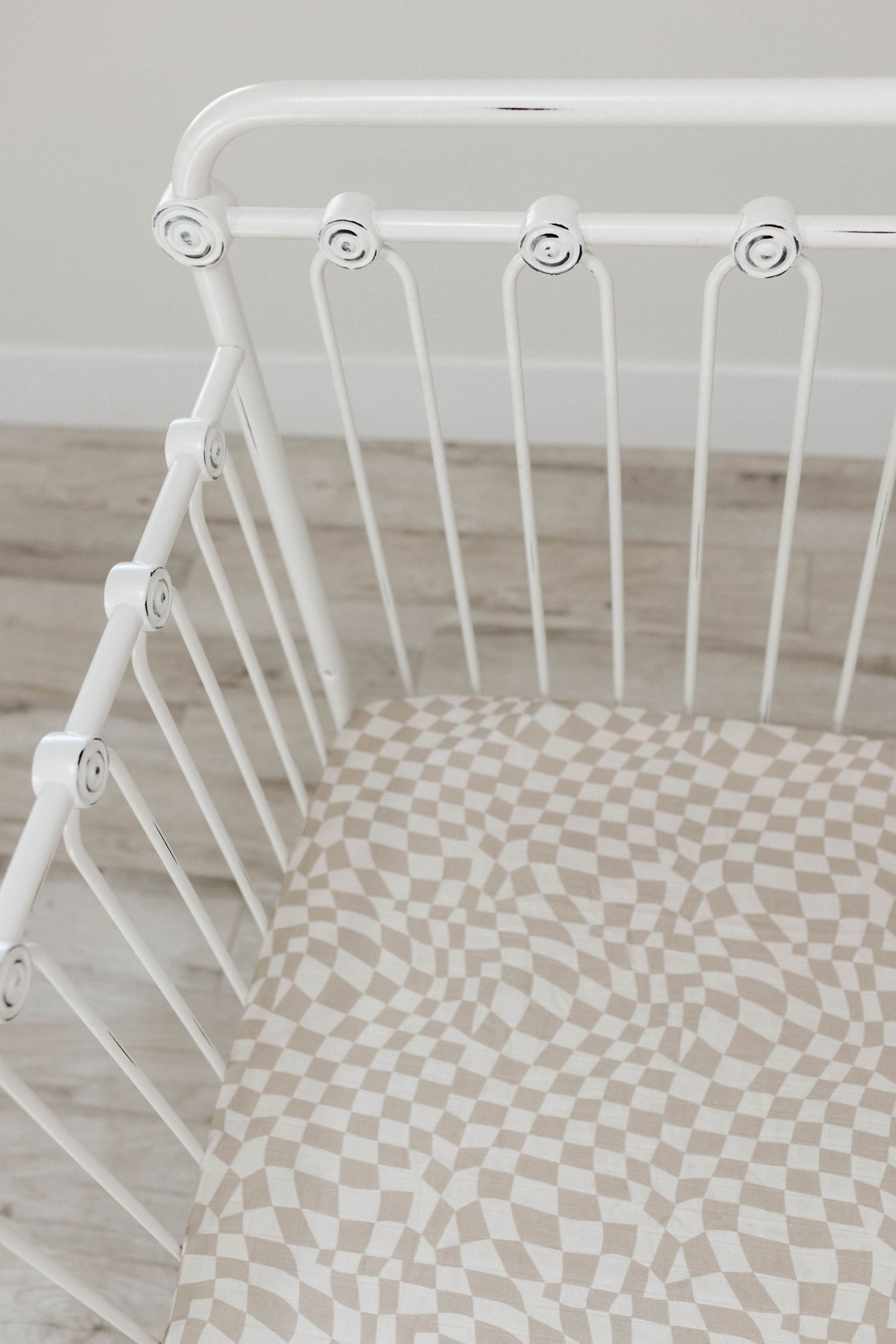 A white metal baby crib with a decorative design on the frame is placed on a light wood floor. The crib mattress, made from soft muslin cotton, is covered with the beige and white *Muslin Crib Sheet | Wavy Checker* sheet by *forever french baby*.