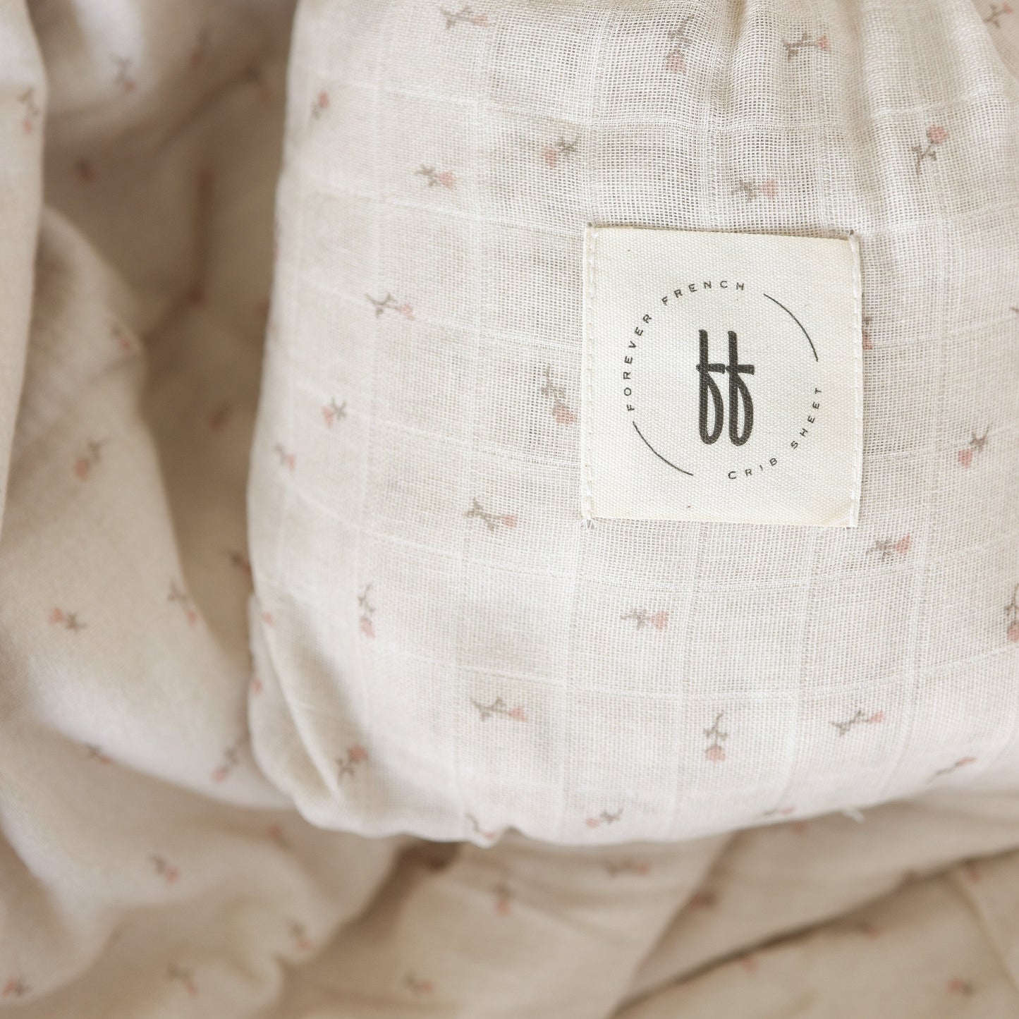 A close-up of the Muslin Crib Sheet | Lucy by forever french baby, featuring soft beige cotton with a pink floral pattern and a stitched “Forever French” label—perfect for an elegant floral nursery.