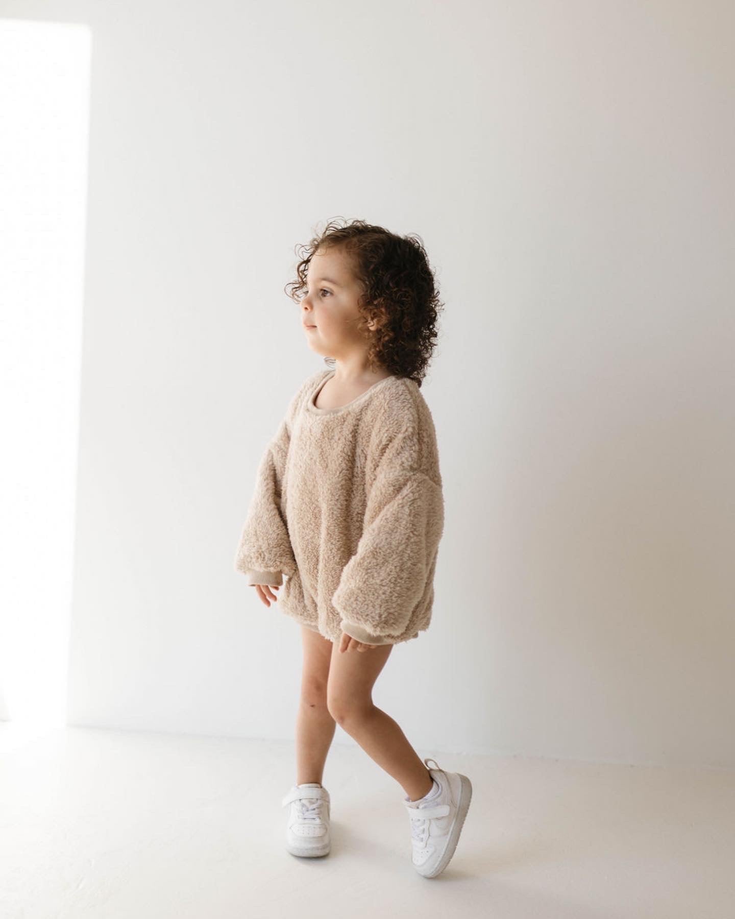 A young child with curly hair stands against a plain white background, wearing an oversized, fuzzy Long Sleeve Romper | Teddy Bear by forever french baby and white sneakers. They are gazing to the left with a relaxed expression as sunlight streams in from the same direction.