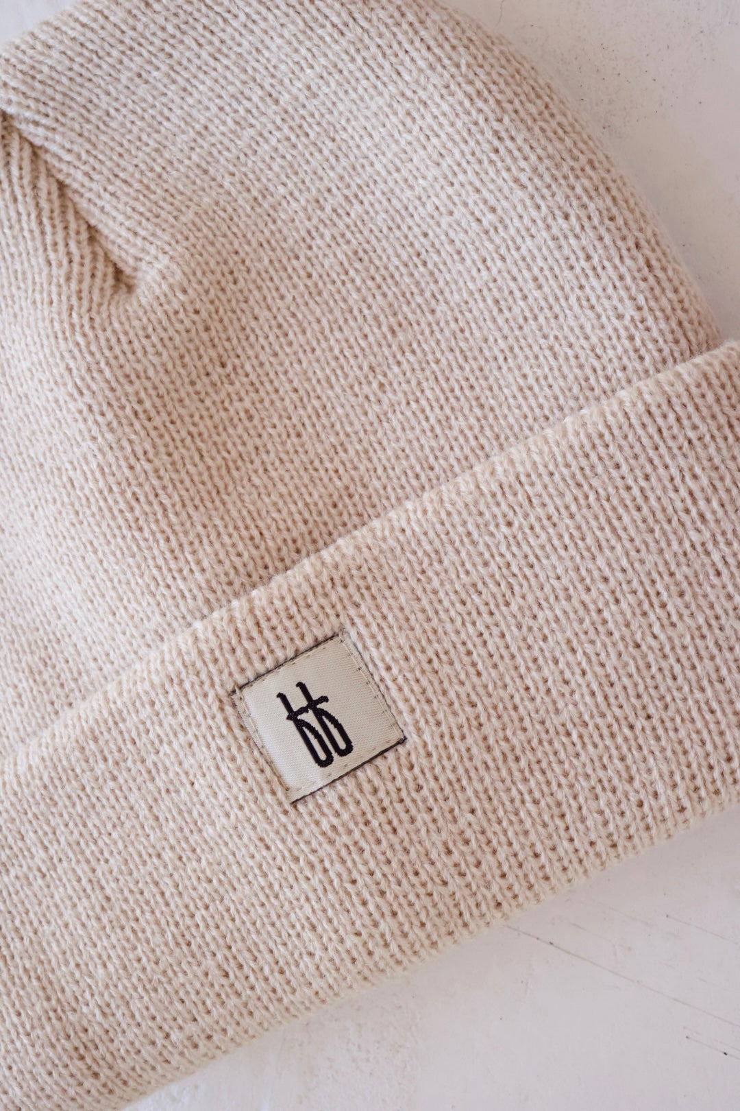 A close-up image of a beige FF Knit Beanie for adults by forever french baby. This knit beanie features a visible fabric tag with a black logo or design. The hat has a folded brim, and the ribbed texture is clearly seen. The background is a light, neutral color.