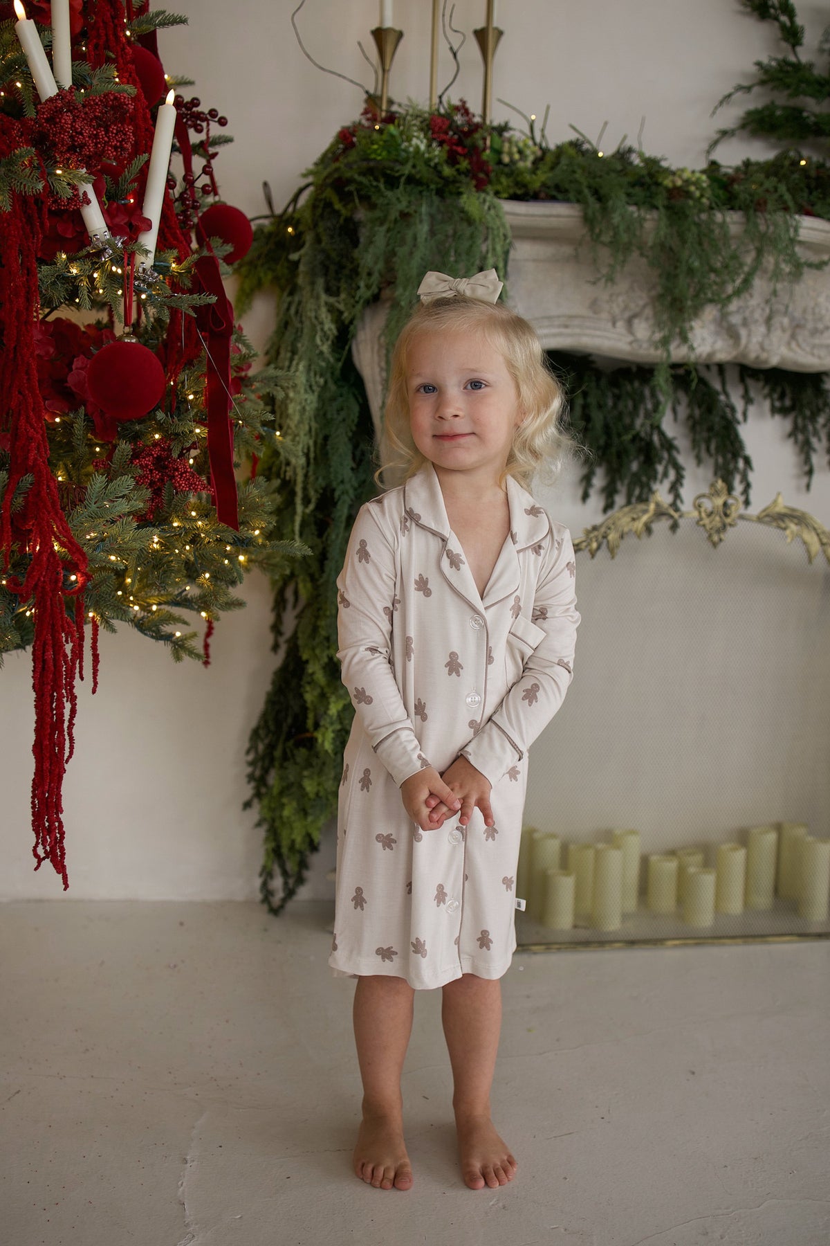 A young child wearing a Bamboo Sleeping Dress | Gingersnap, a breathable, long-sleeved button-up nightgown with a repeating gingerbread man pattern.