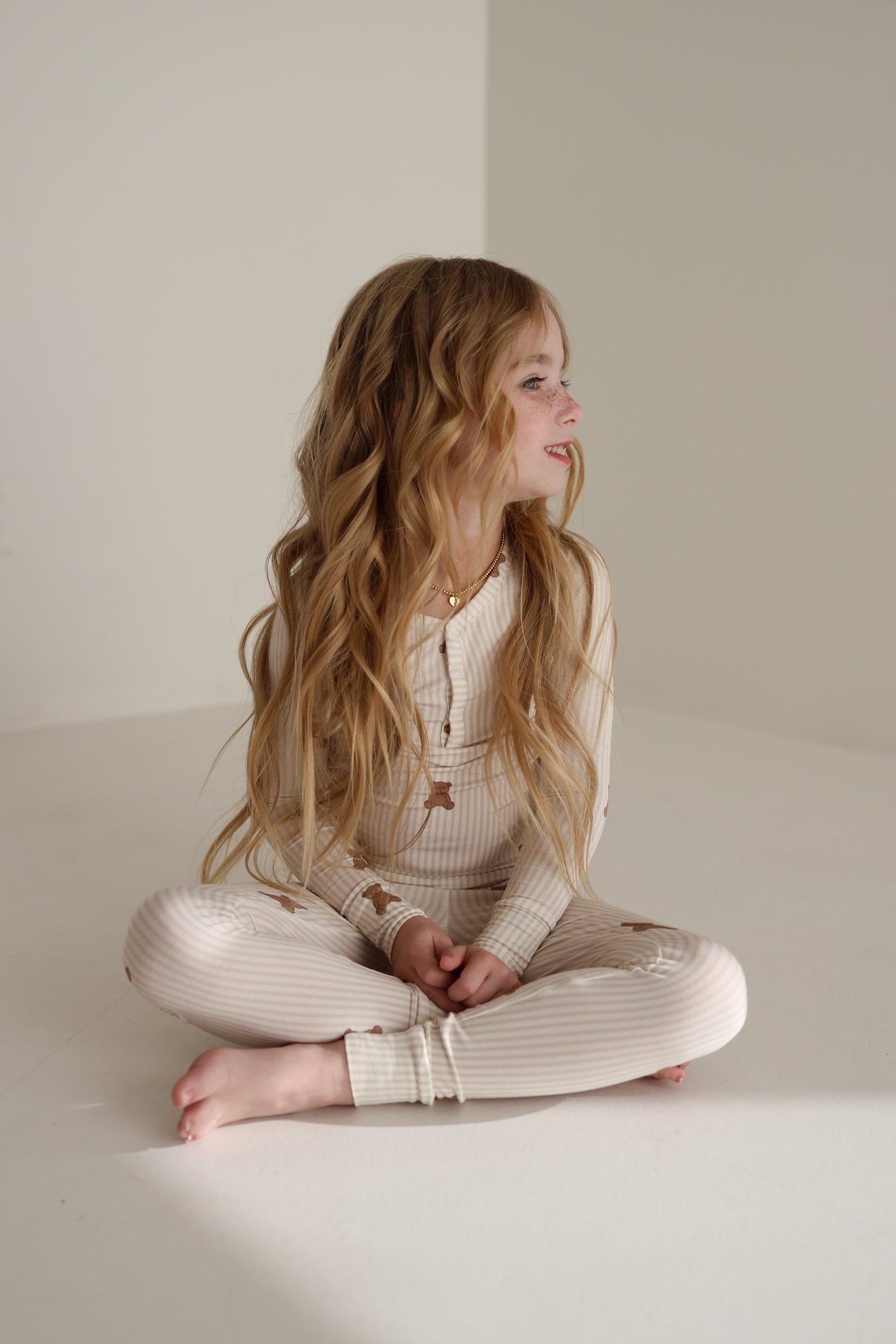 A young girl with long wavy blonde hair sits cross-legged on the floor, wearing forever french baby’s Bamboo Two Piece Pajamas | Teddy. She looks to her right against a plain, neutral background.