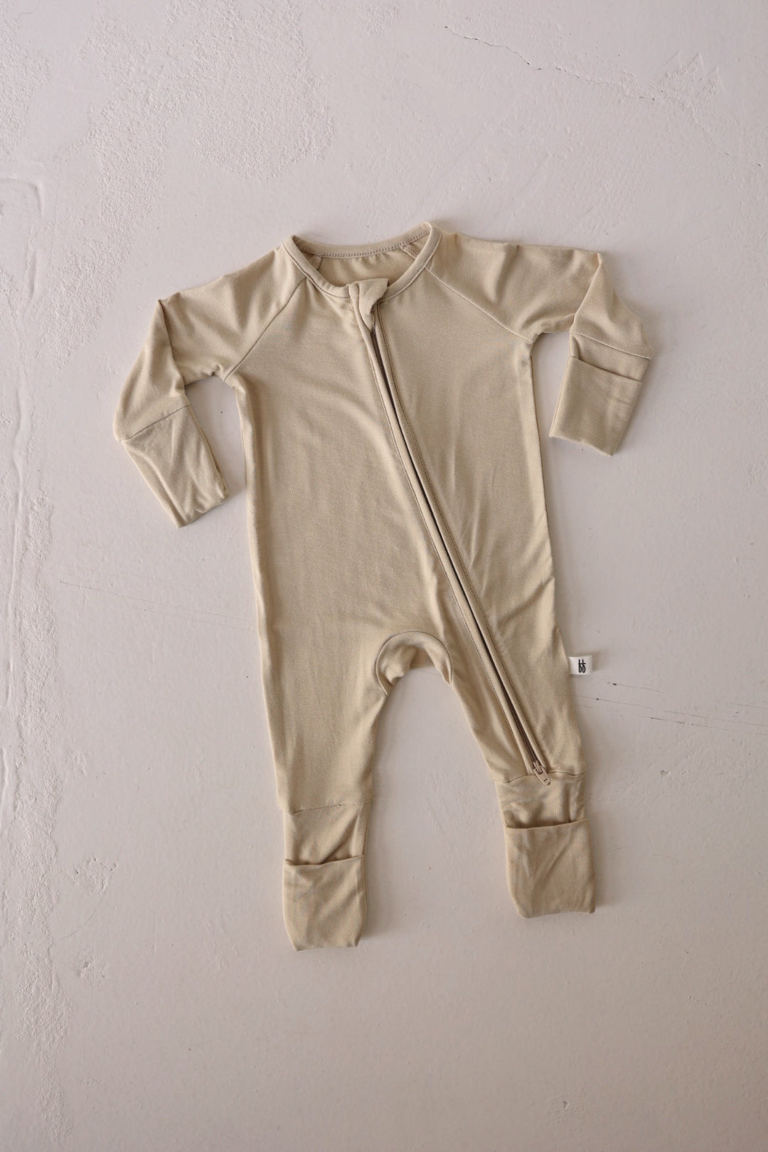 The Bamboo Zip Pajamas in Limestone, from forever french baby, are laid flat on a white textured surface. This beige onesie features long sleeves, long legs, foldable cuffs, and an asymmetrical zipper running from the neck to one leg. Crafted as hypo-allergenic clothing, it's gentle on delicate skin for a peaceful night's rest.