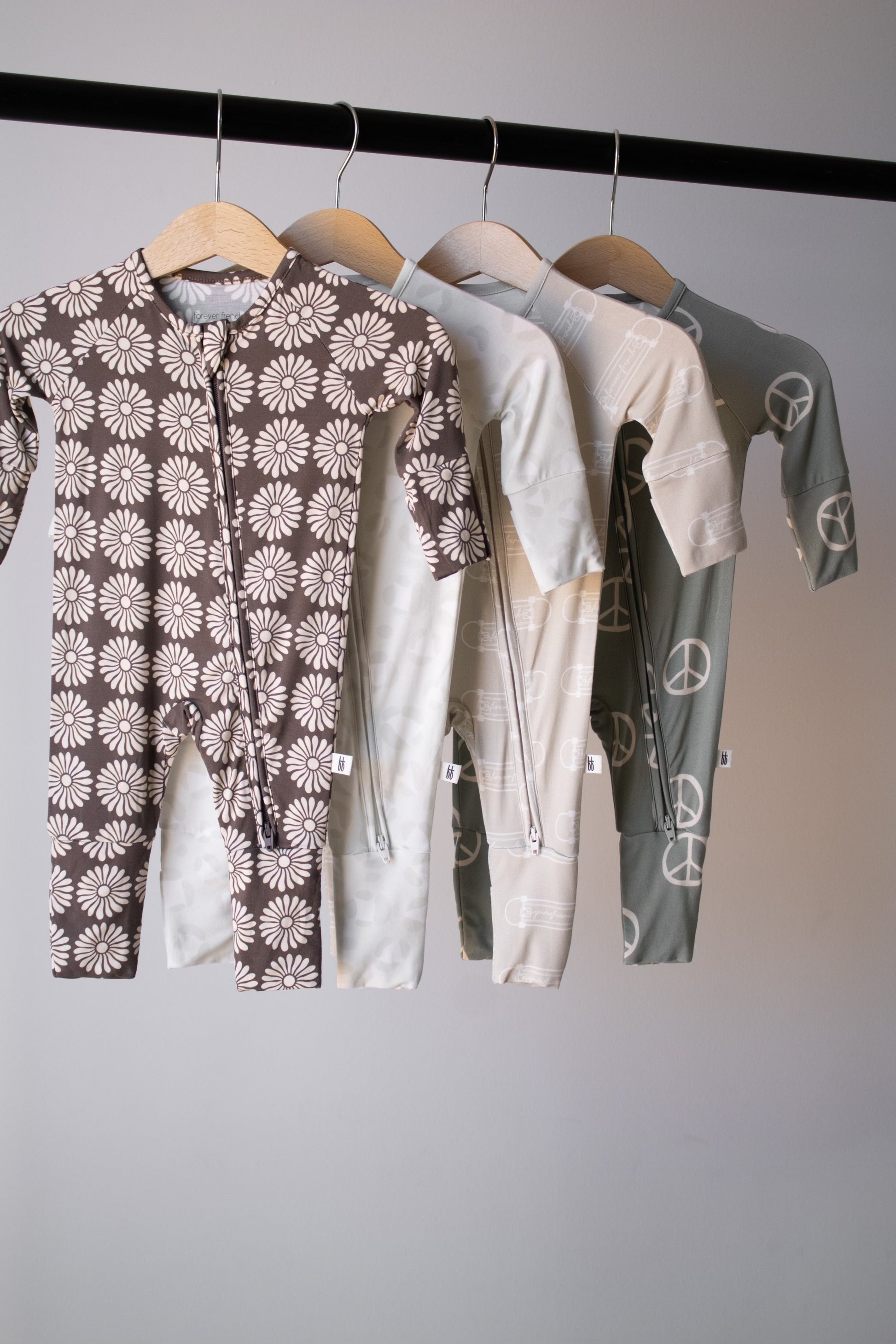 Four Bamboo Zip Pajamas | Retro Floral by forever french baby, featuring daisy and peace sign patterns, hang on wooden hangers from a black rod against a plain light gray background.