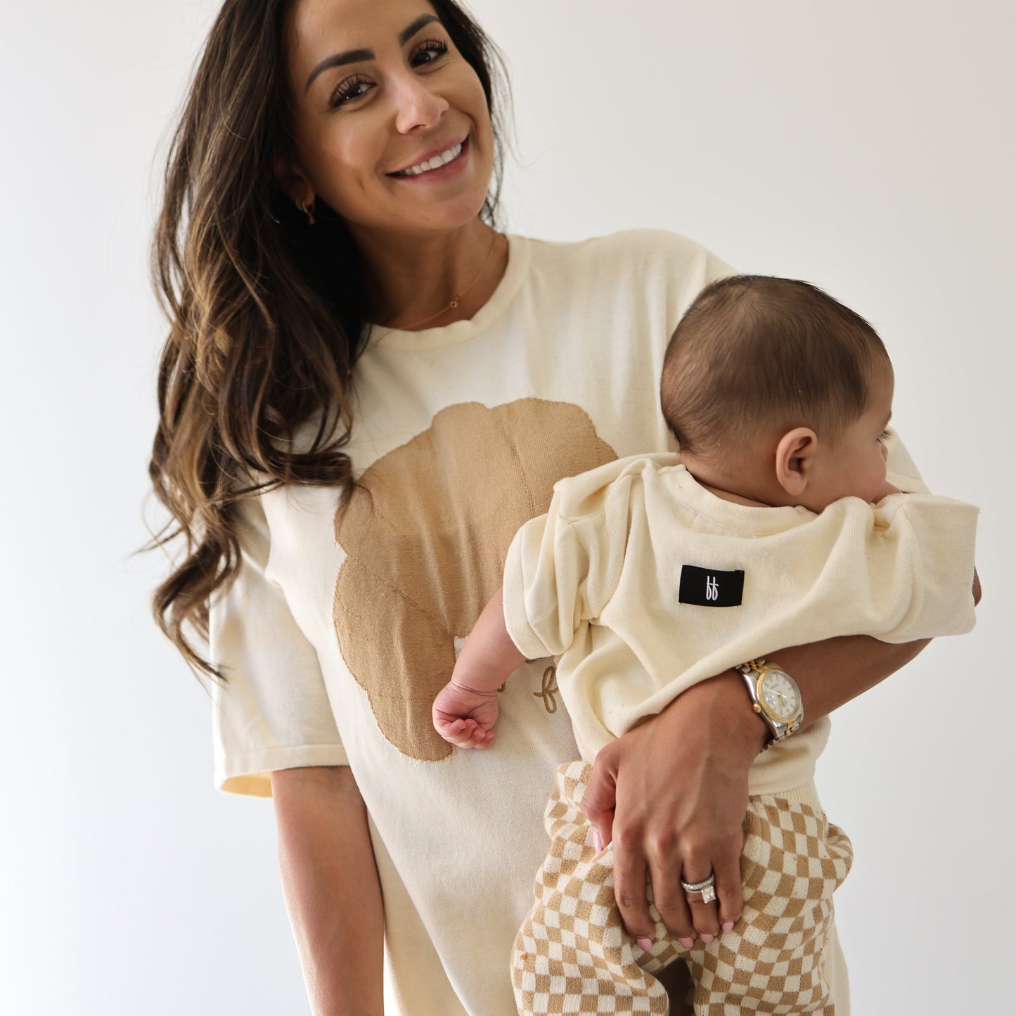 A smiling woman with long dark hair holds a baby dressed in the forever french baby Knit Pant Set | Croissants, featuring a beige top and checkered pants, against a plain light background.