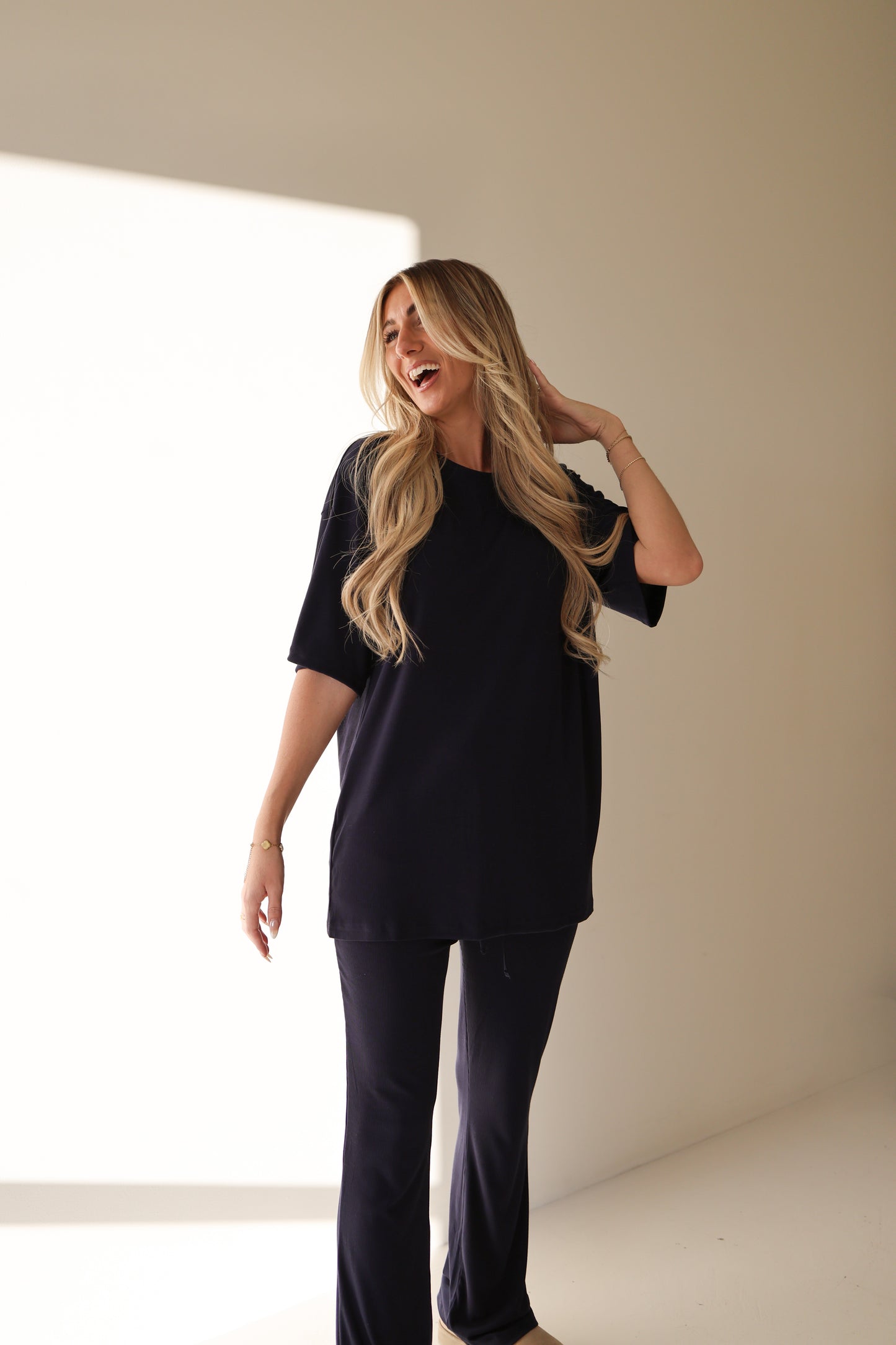 A smiling woman with long, wavy blonde hair touches her hair while wearing the forever french baby Short Sleeve Bamboo Women's Lounge Set in Blueberry Ribbed, standing in a brightly lit indoor space with a shadow on the wall.