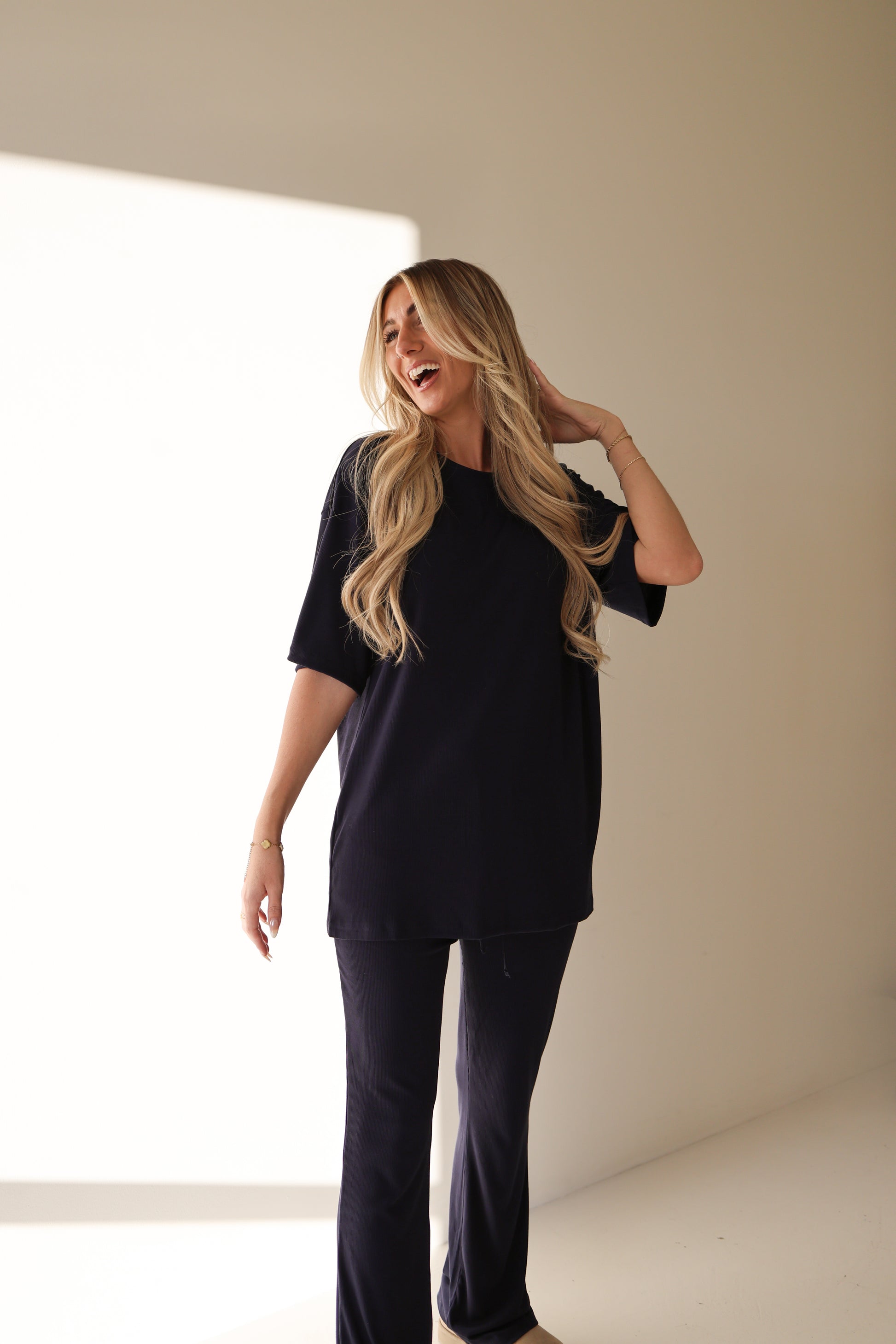 A smiling woman with long, wavy blonde hair touches her hair while wearing the forever french baby Short Sleeve Bamboo Women's Lounge Set in Blueberry Ribbed, standing in a brightly lit indoor space with a shadow on the wall.