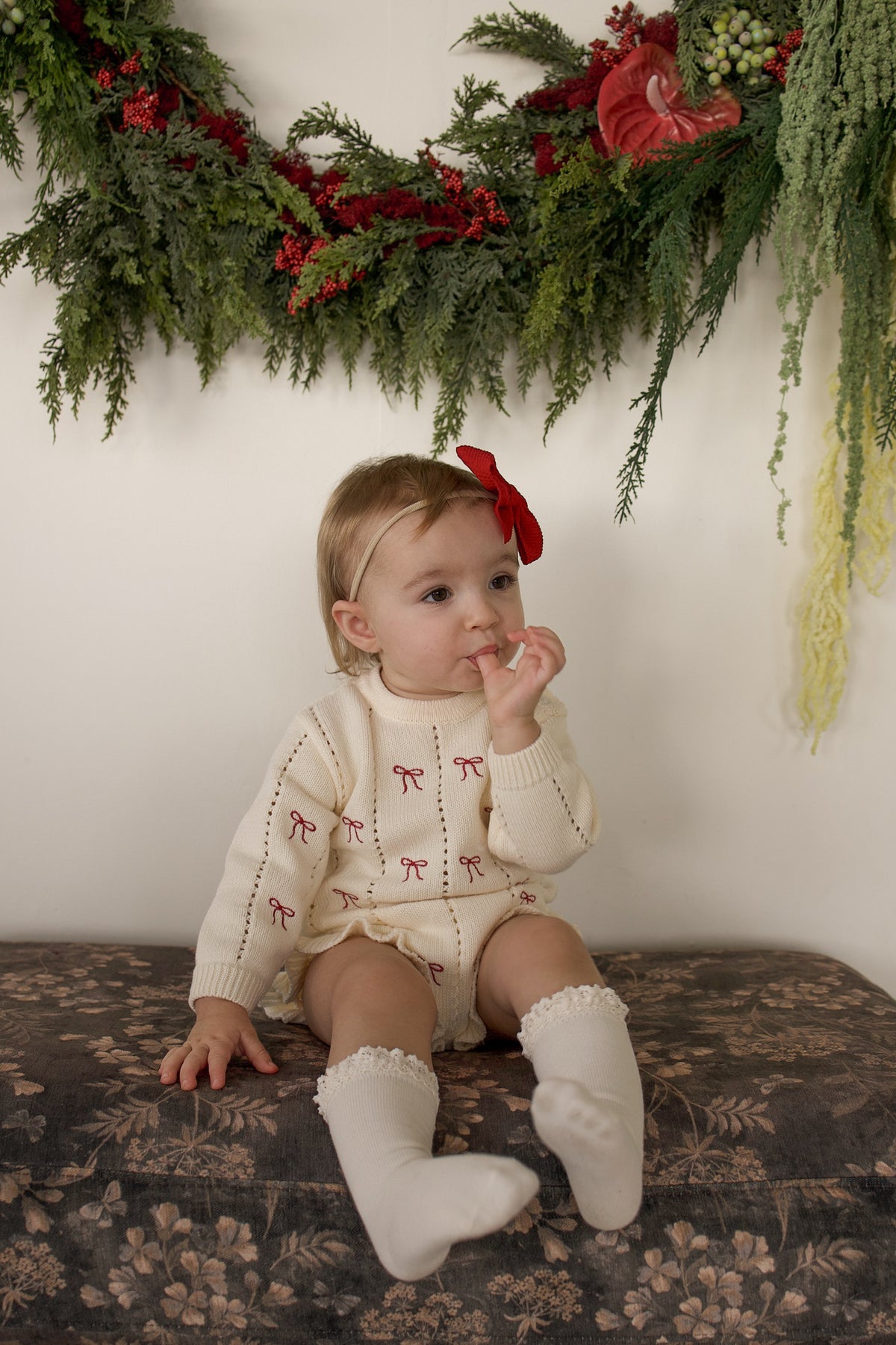 A toddler wearing a Knit Ruffle Romper | Candy Cane with small red bows and matching ruffled socks sits on a patterned bench.
